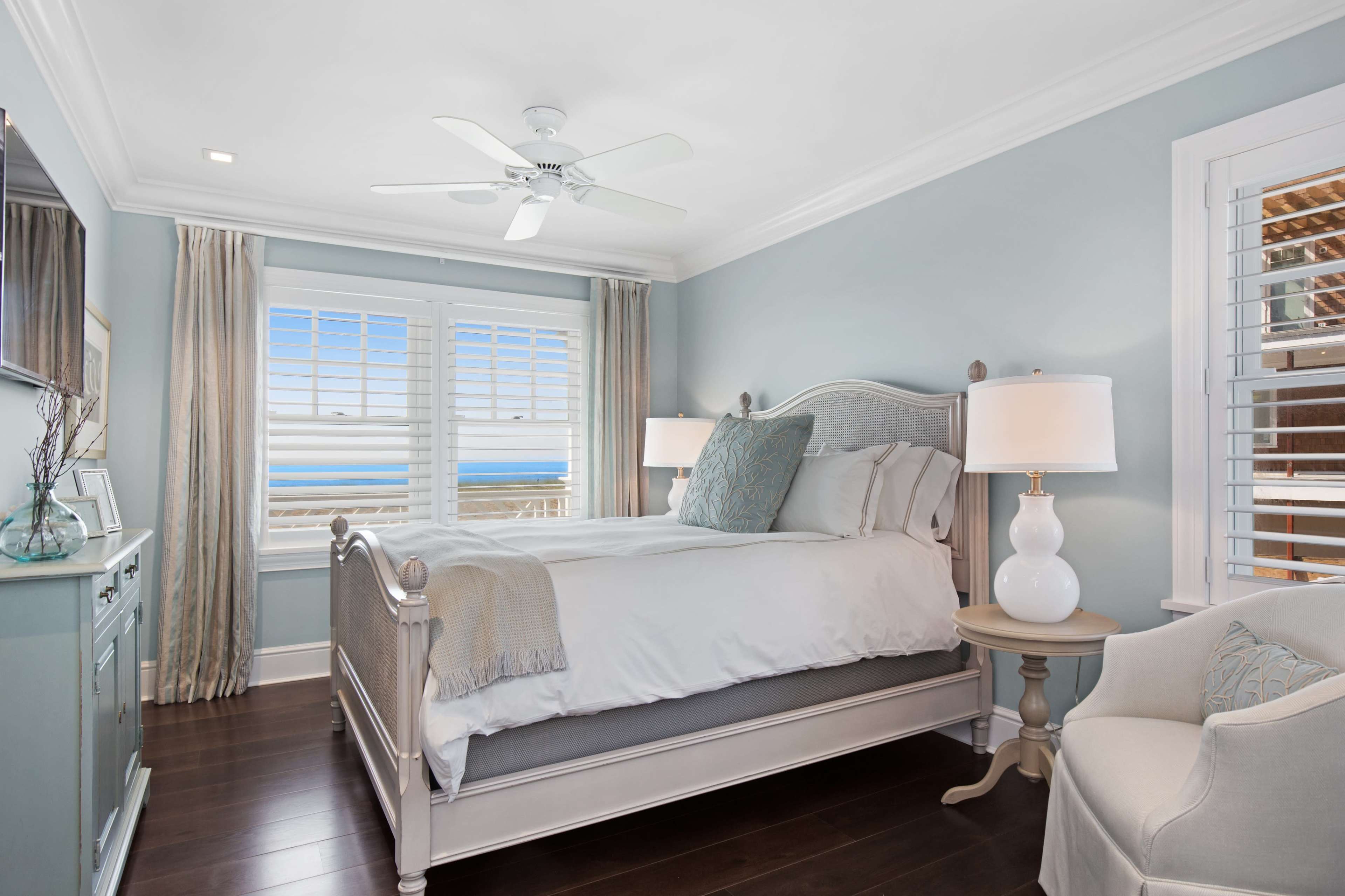 A decorated bedroom features a light blue wall, a white bed with a gray blanket, and a window showing an ocean view.