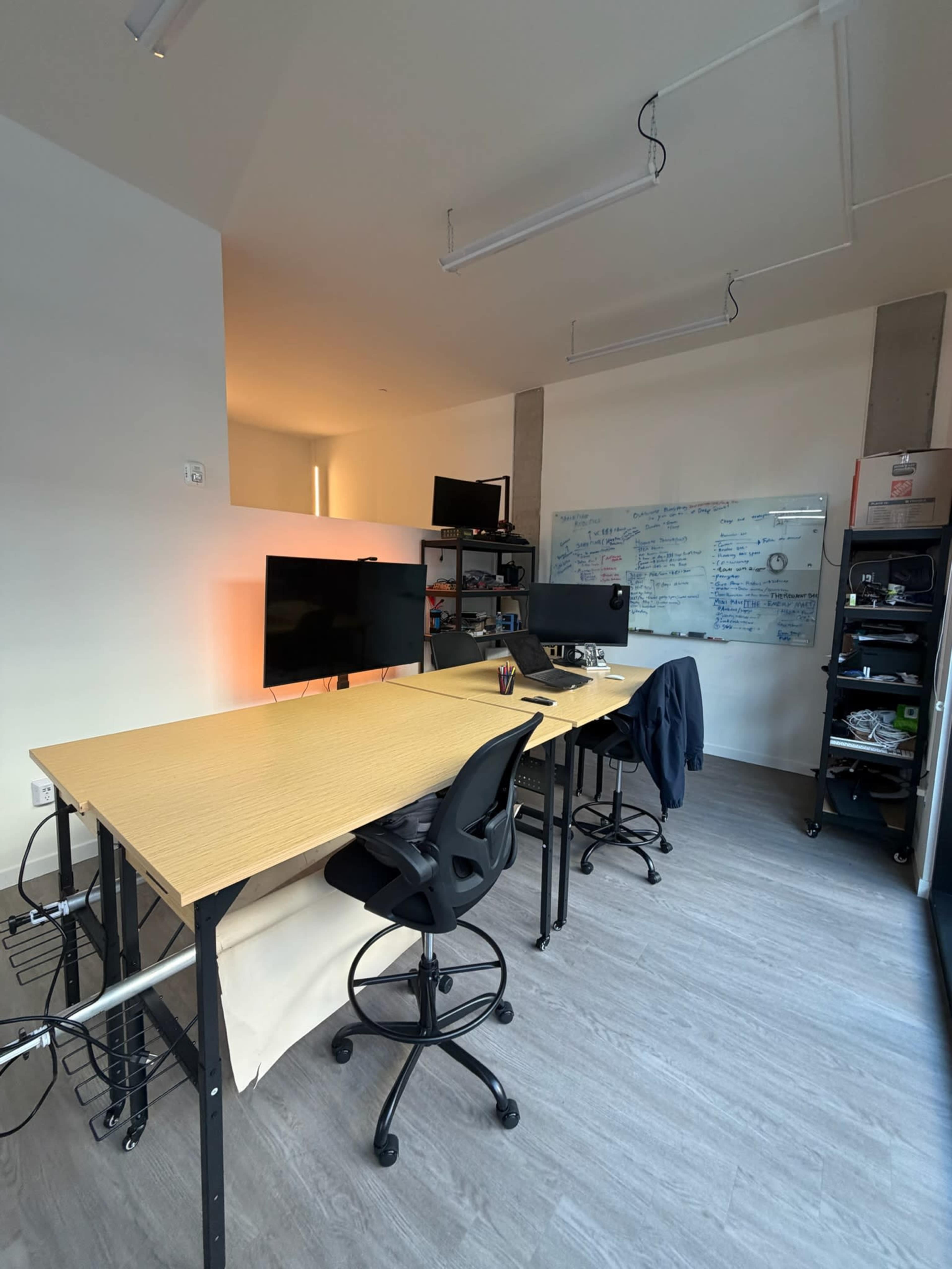 The image shows a modern office setup with three height-adjustable desks, two monitors, a whiteboard covered in writing, and a shelf filled with supplies.