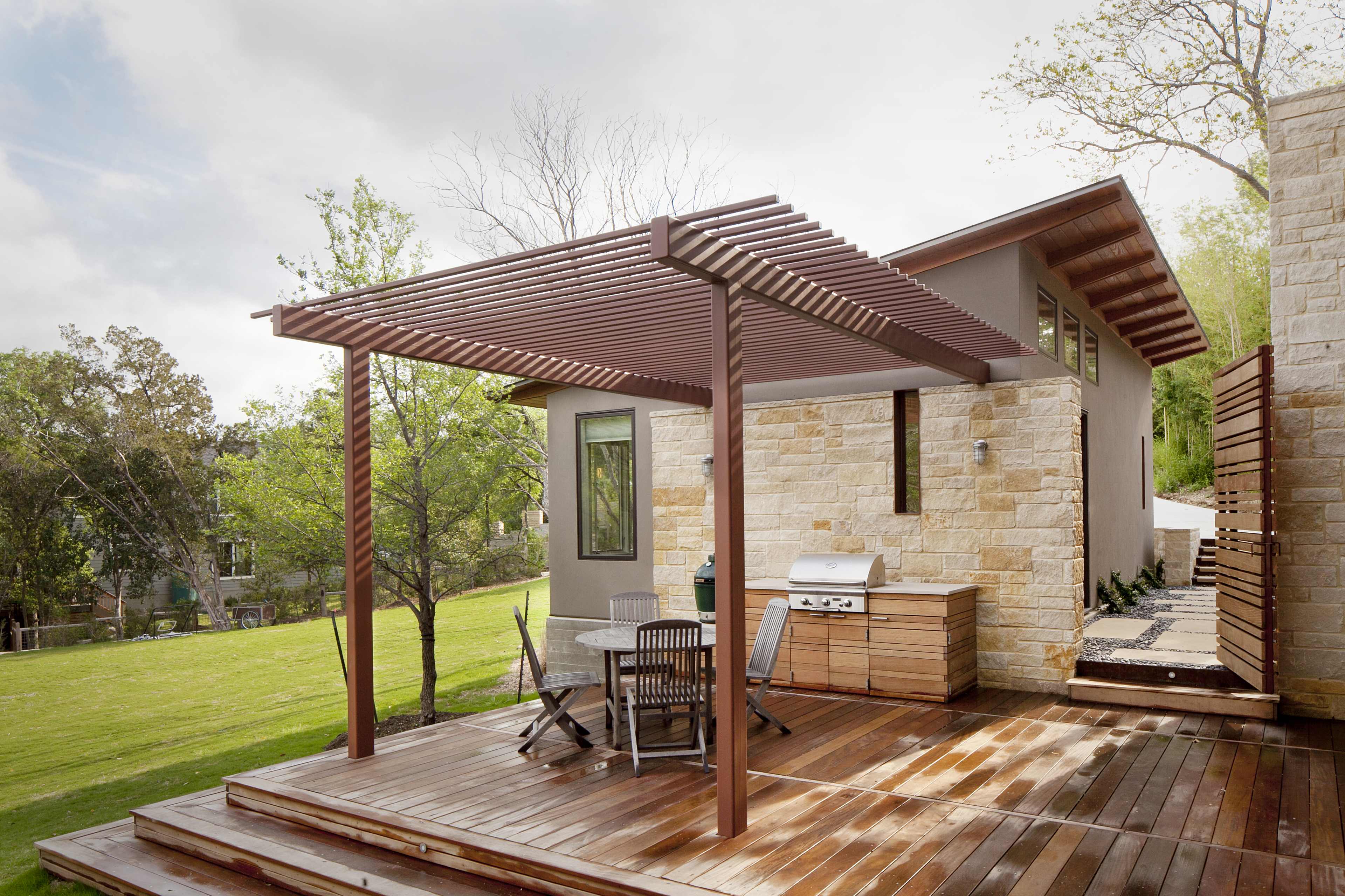 The image shows a modern wooden deck with a pergola, a grill area, and a stone-and-wood exterior of a house surrounded by greenery.