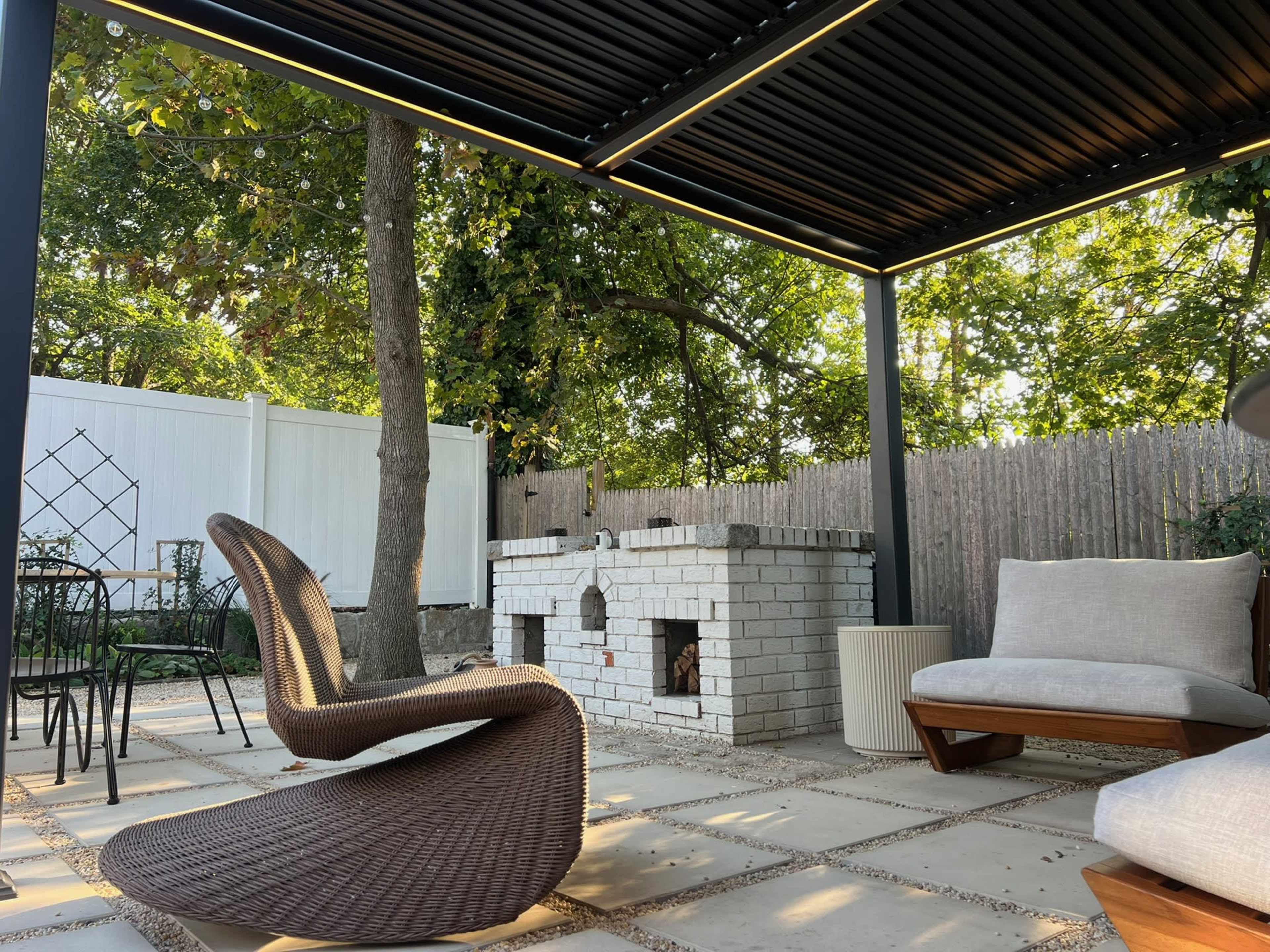 A shaded outdoor patio features a modern lounge chair, a brick outdoor fireplace, and surrounding greenery.