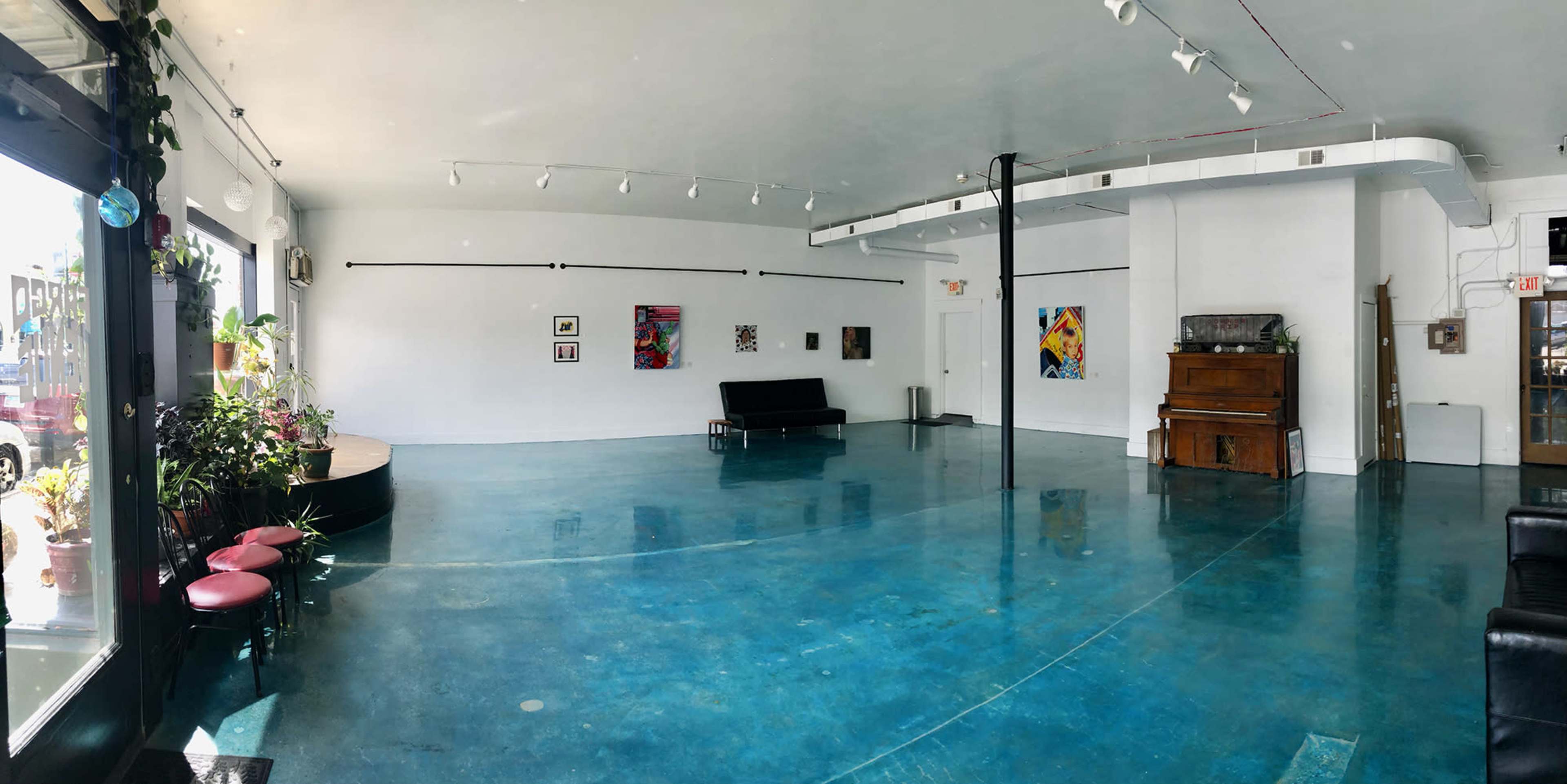 The image shows a spacious, brightly lit gallery with a polished blue concrete floor, featuring a few pieces of art on the white walls and some seating arrangements.