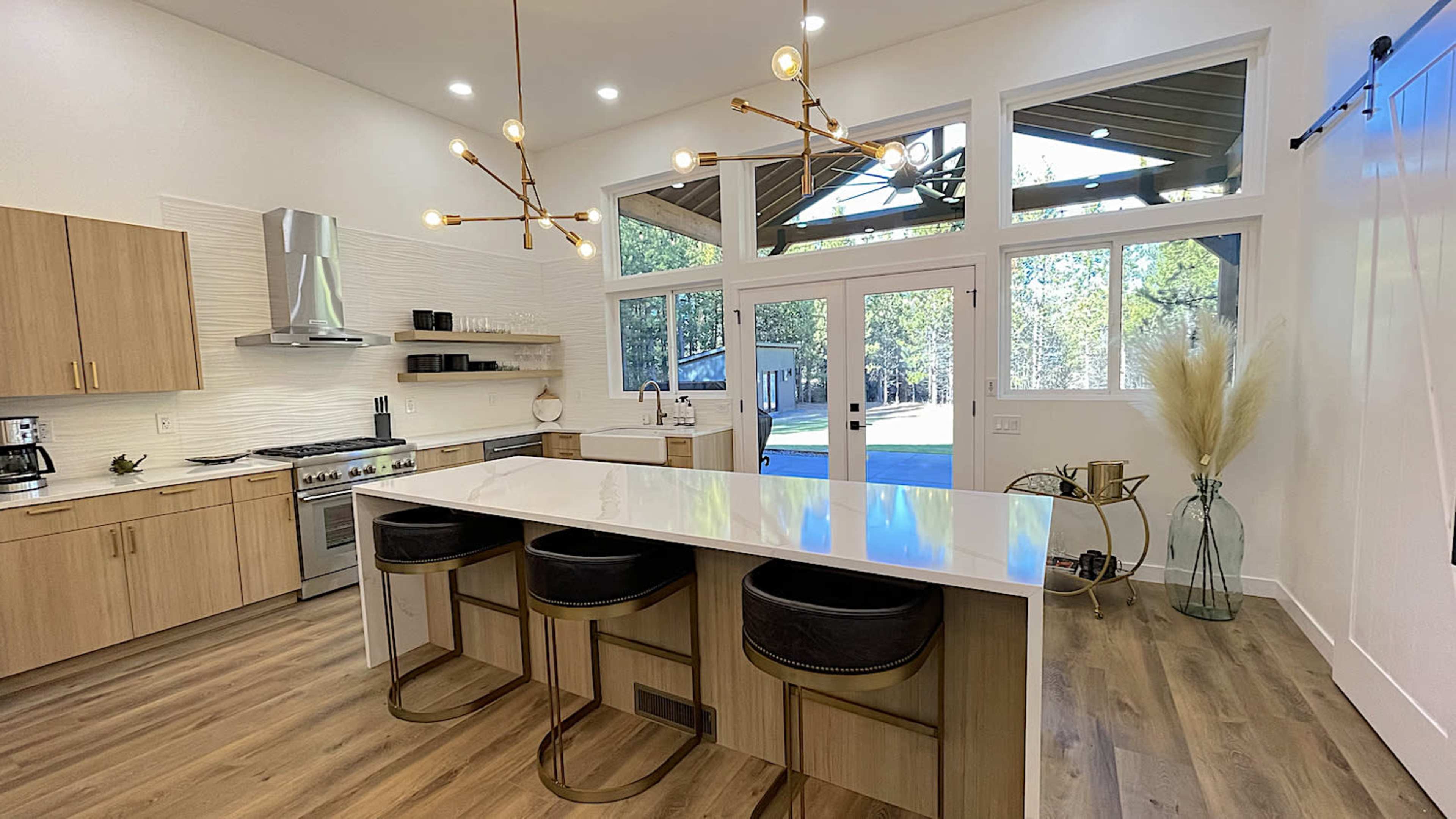 A modern kitchen with a large island fitted with black stools, stainless steel appliances, and large windows overlooking a wooded area.