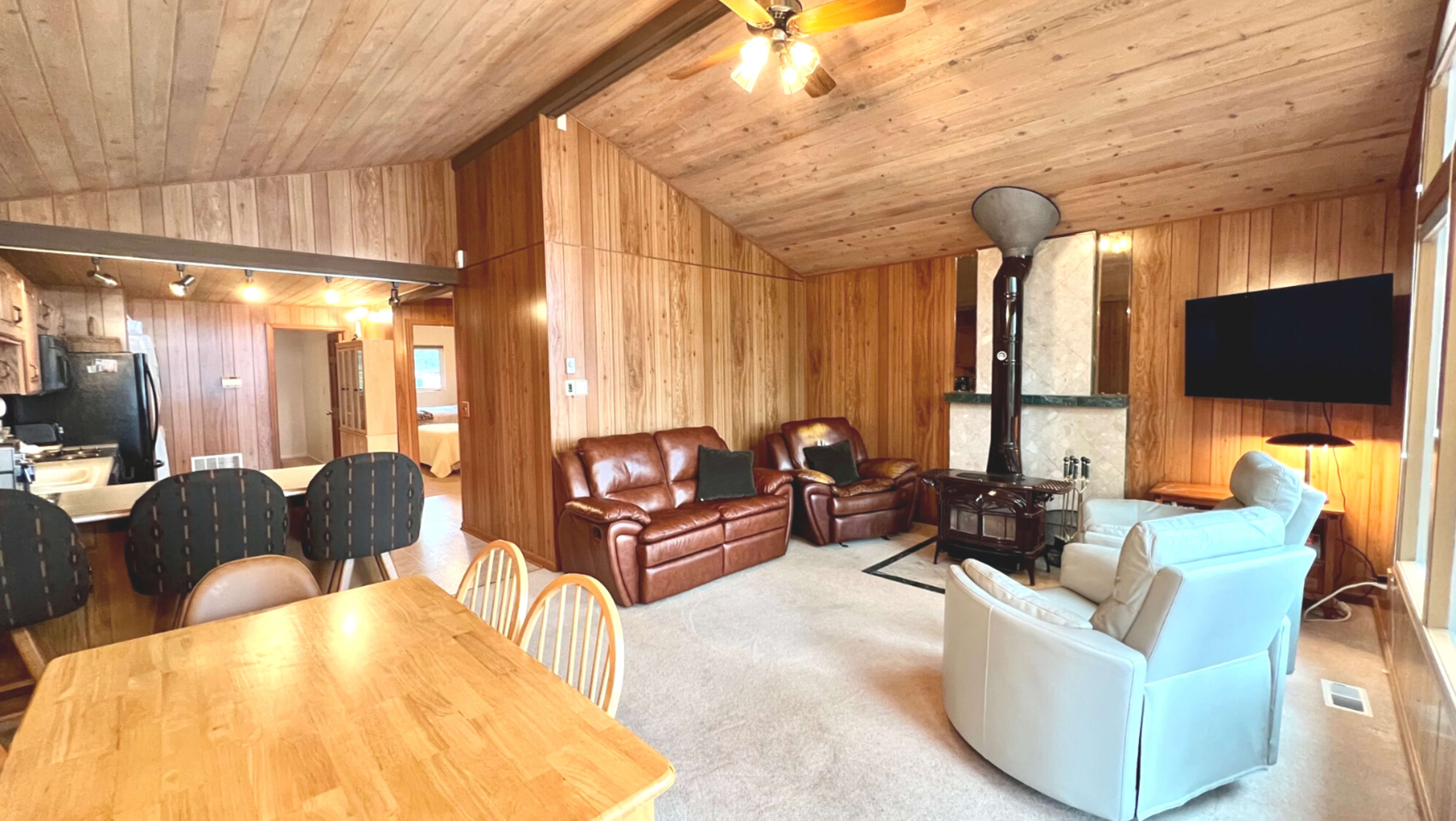 A cozy living area featuring wooden walls, a dining table with chairs, leather sofas, and a corner fireplace.