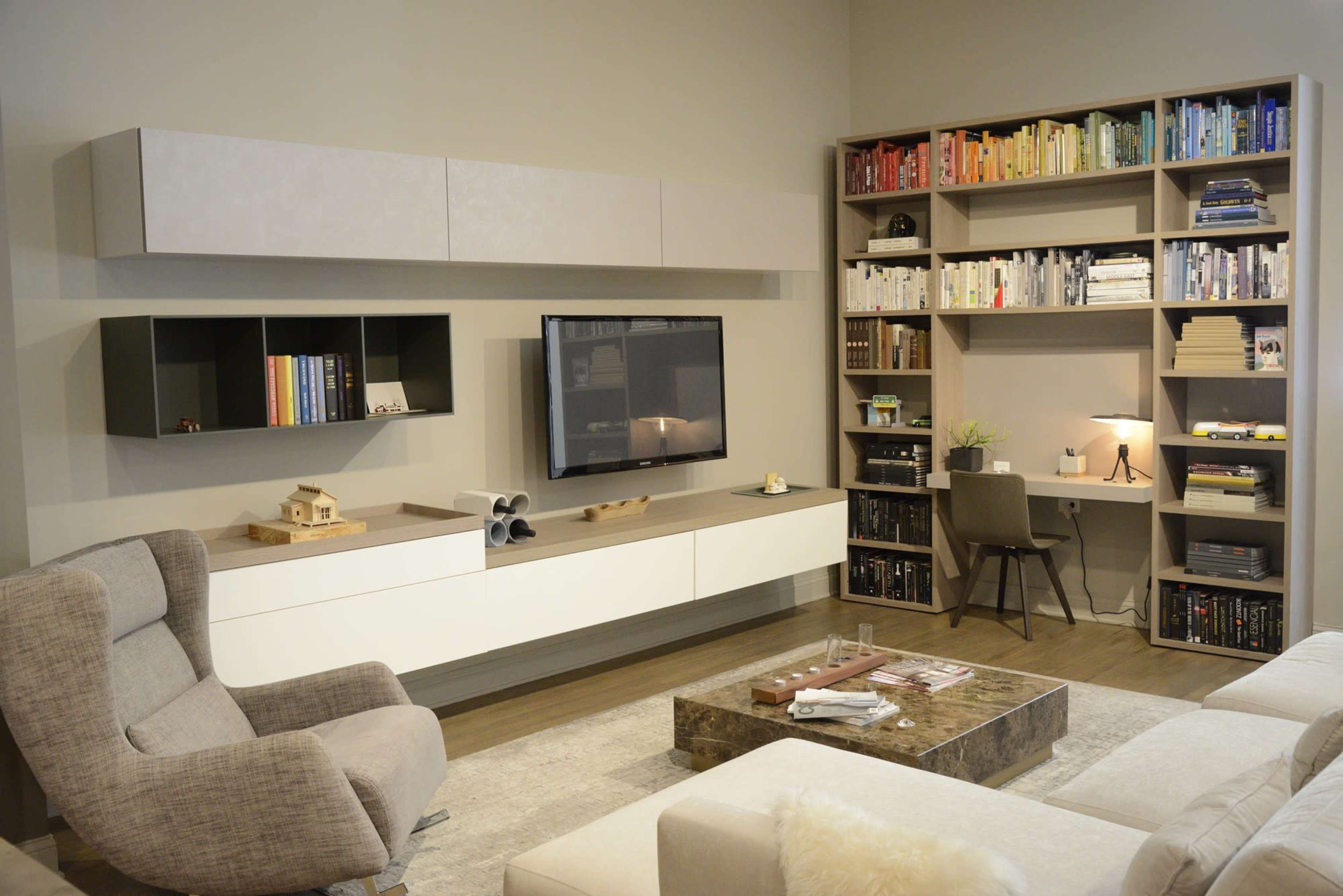A modern living room features a light-colored wall unit with a TV, bookshelves filled with books, a cozy armchair, and a coffee table on a rug.