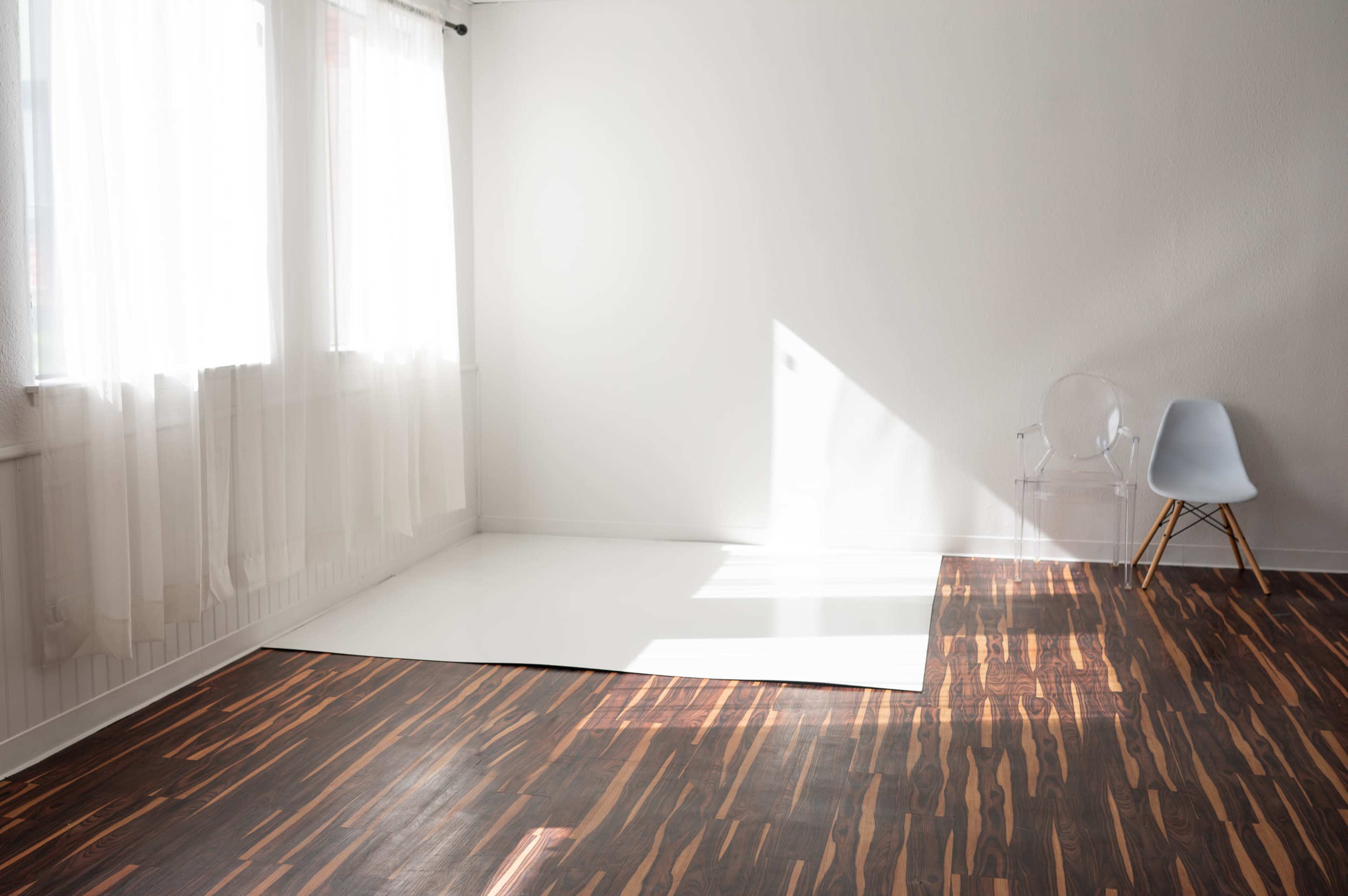 A bright, minimalist room features a blend of light and dark wood flooring, white walls, large windows with sheer curtains, and a transparent chair beside a white area.