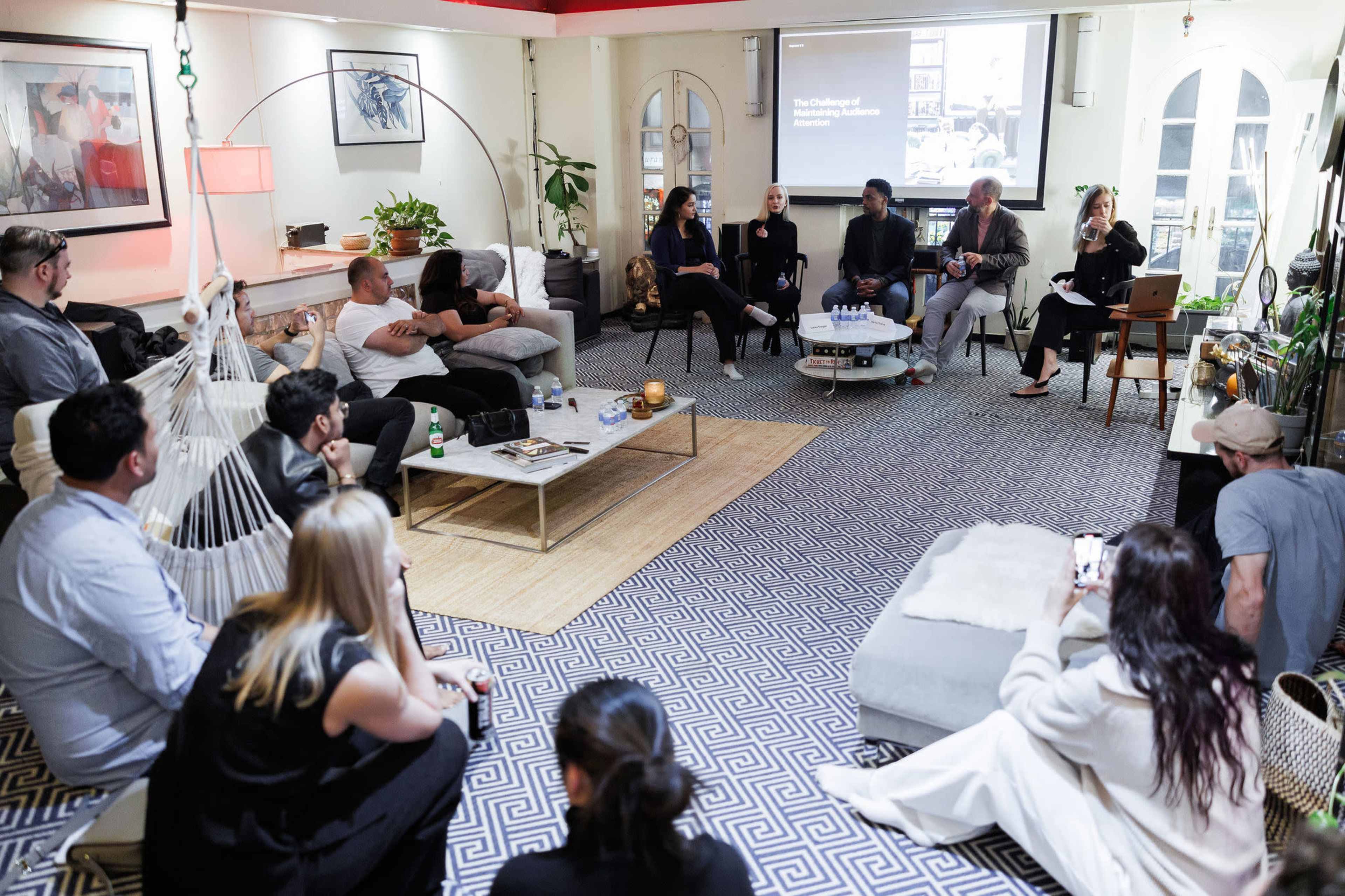 A group of people is seated in a cozy room, engaged in a panel discussion while others listen attentively.