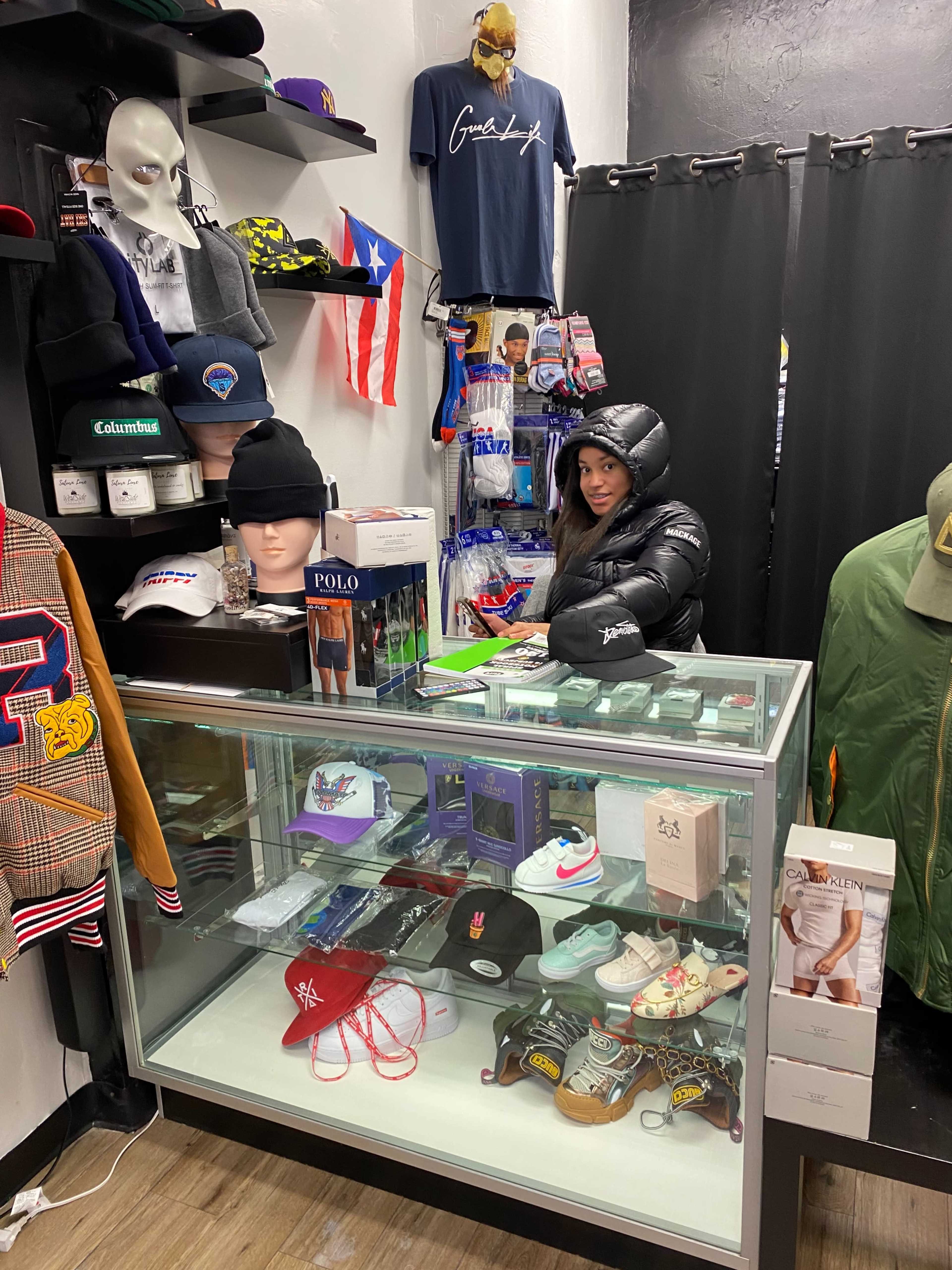 A woman stands behind a glass display case filled with various hats, sneakers, and clothing items in a retail store.
