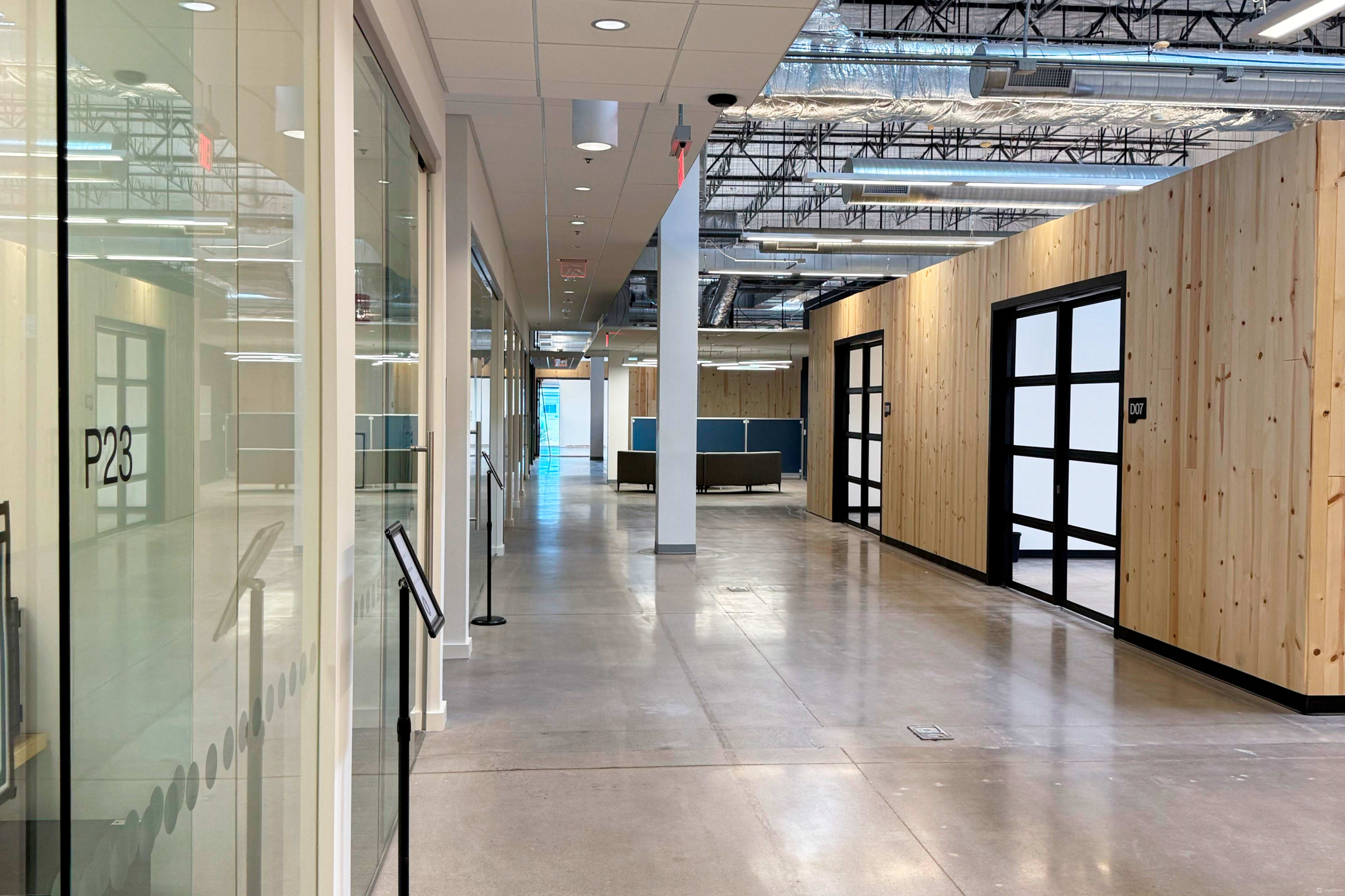 The image shows a modern hallway with glass office doors on one side and wooden paneling on the opposite side, leading to a brighter area at the end.