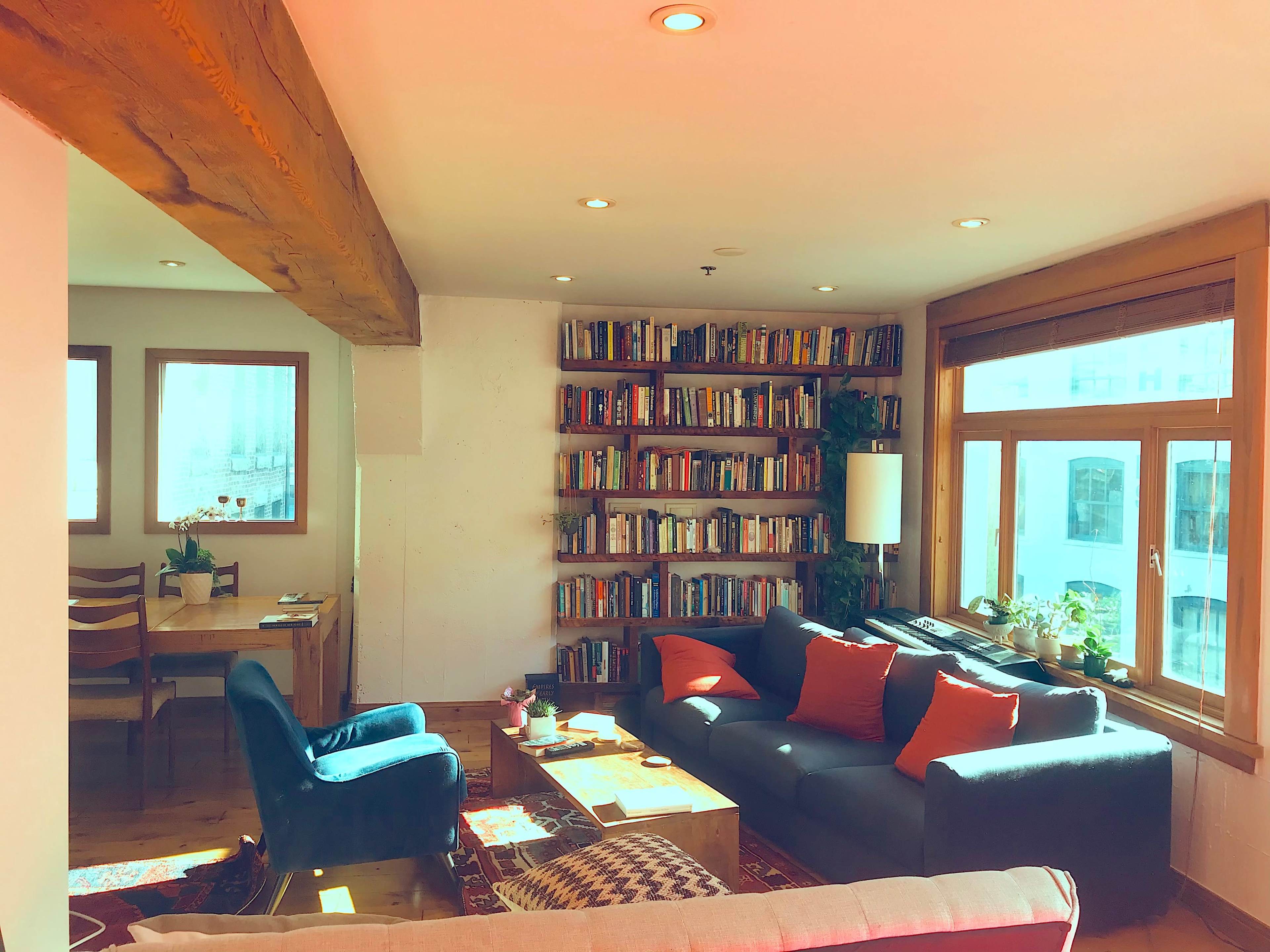 A cozy living room with a bookshelf filled with books, a sofa, and large windows providing natural light.