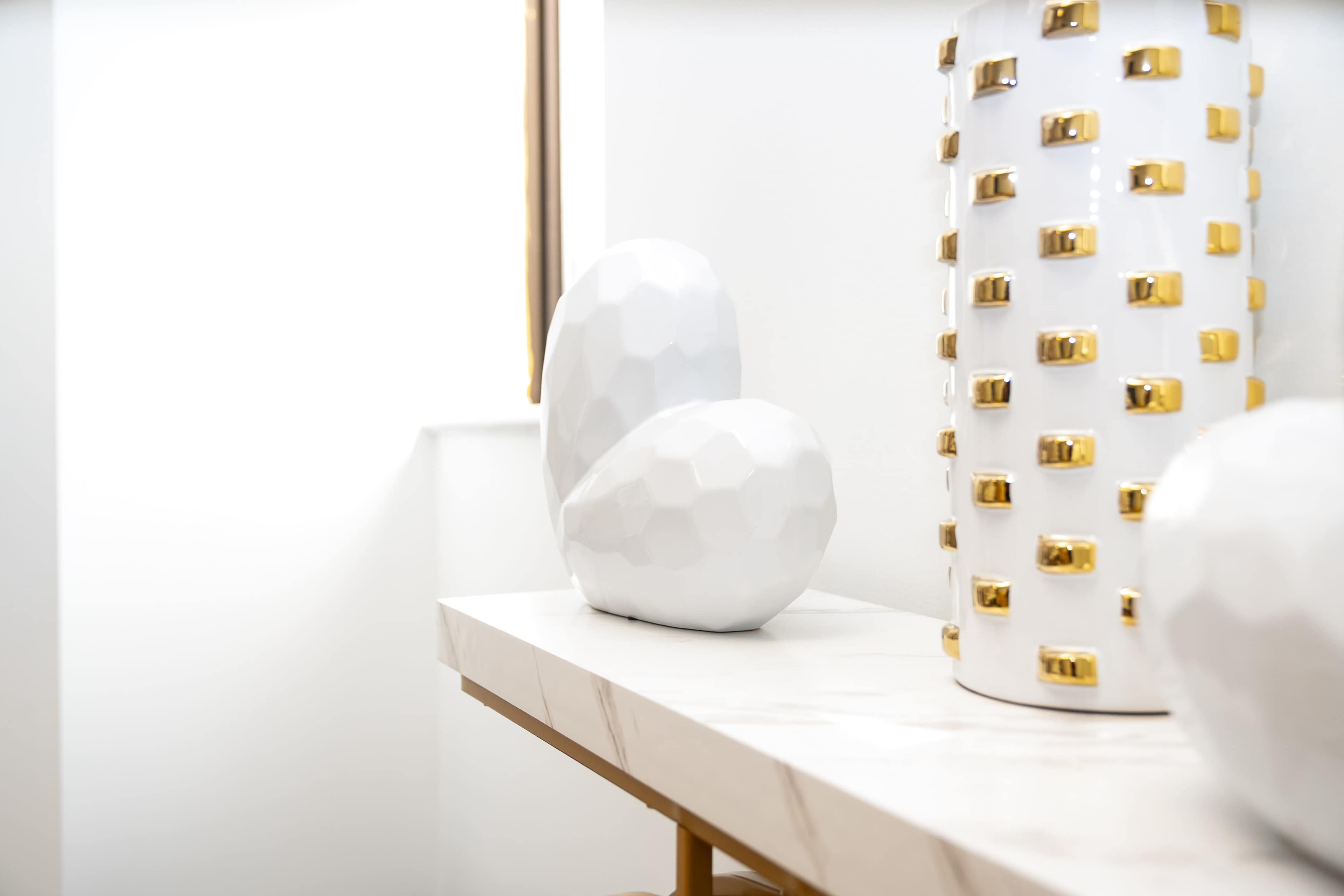 A white marble table with a geometric white vase and a gold-accented cylindrical vase displayed on it.