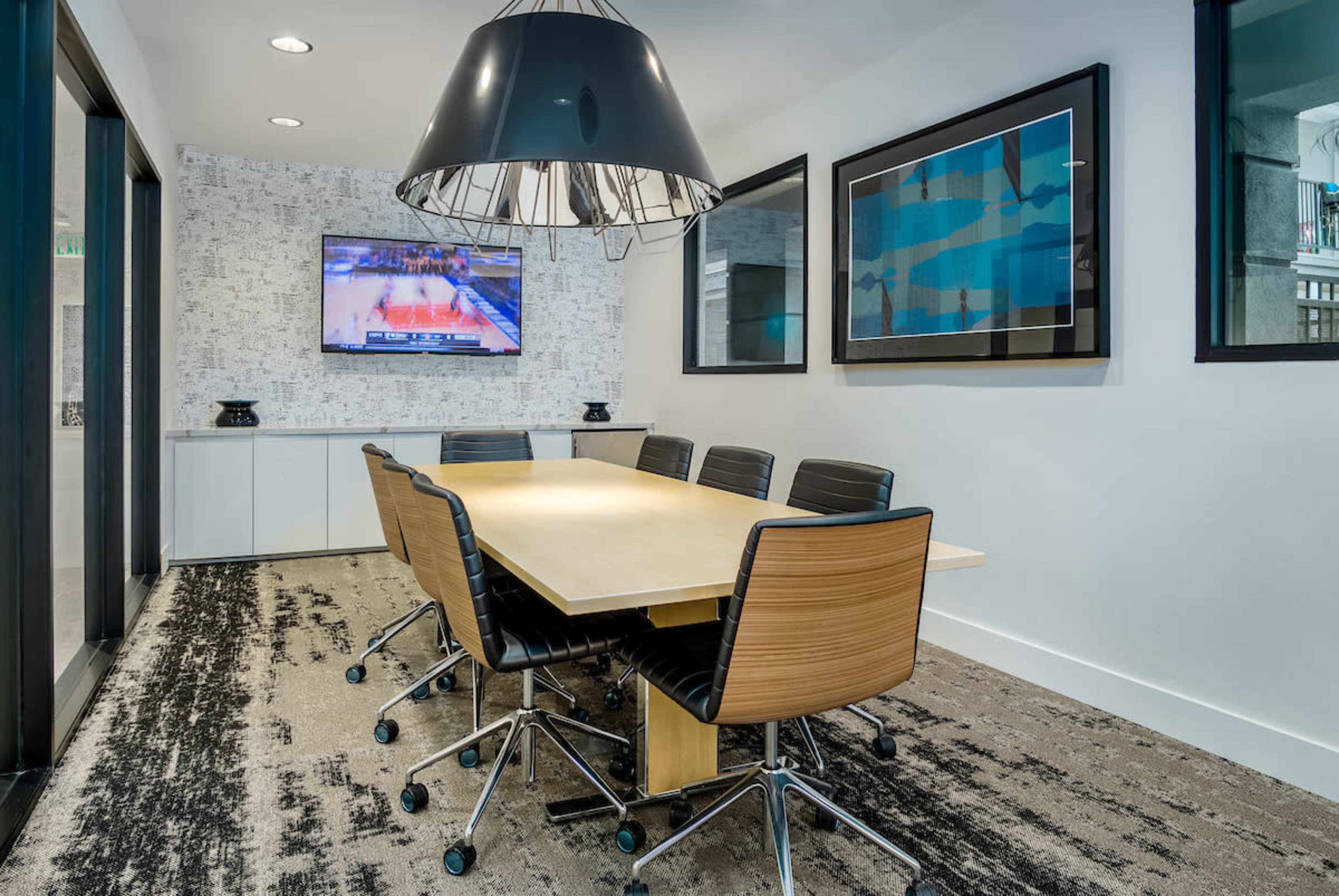 A modern conference room features a large wooden table surrounded by black chairs, with a television displaying a basketball game on the wall.