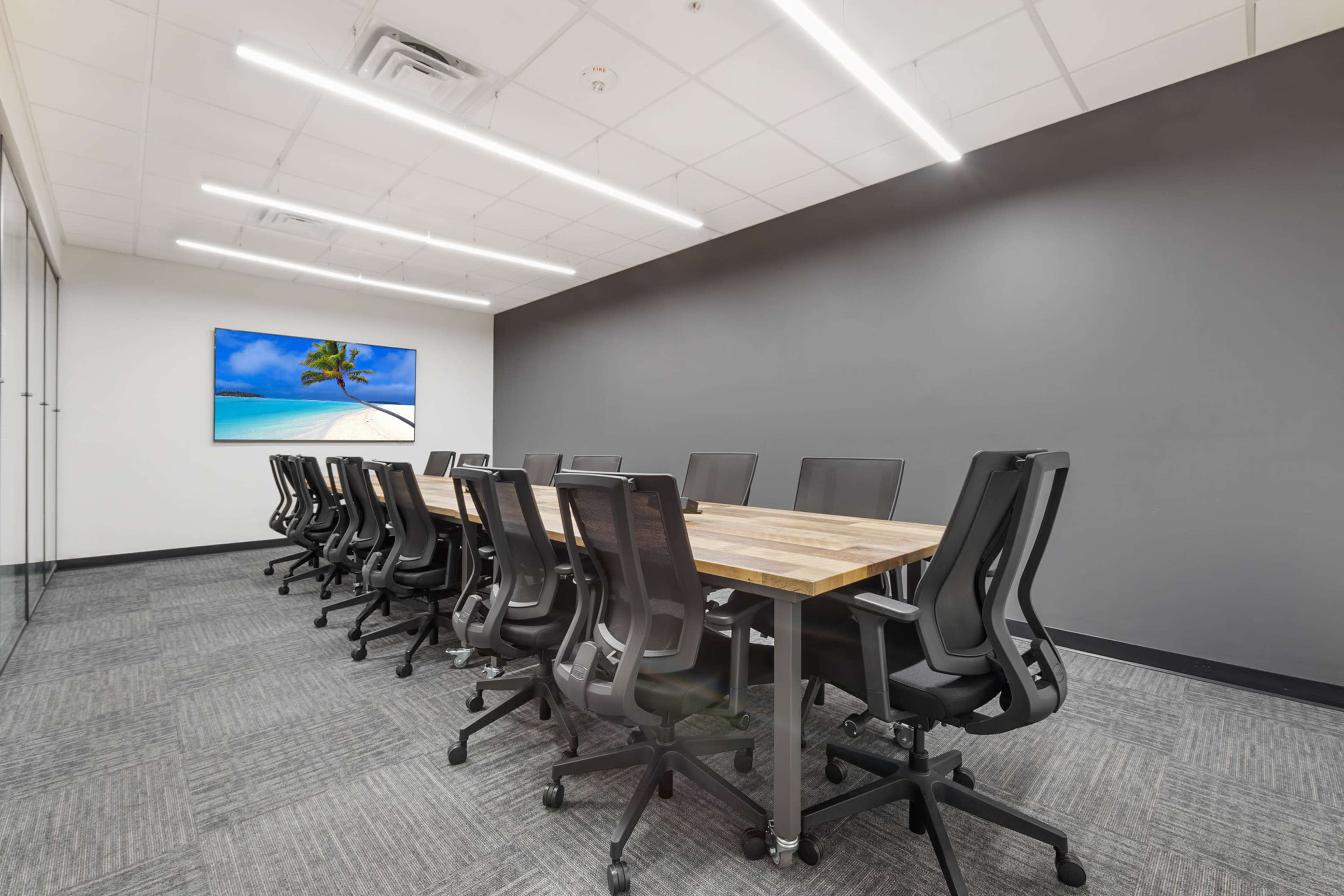 A conference room features a long wooden table surrounded by ergonomic black chairs, with a large screen displaying a beach scene on the wall.