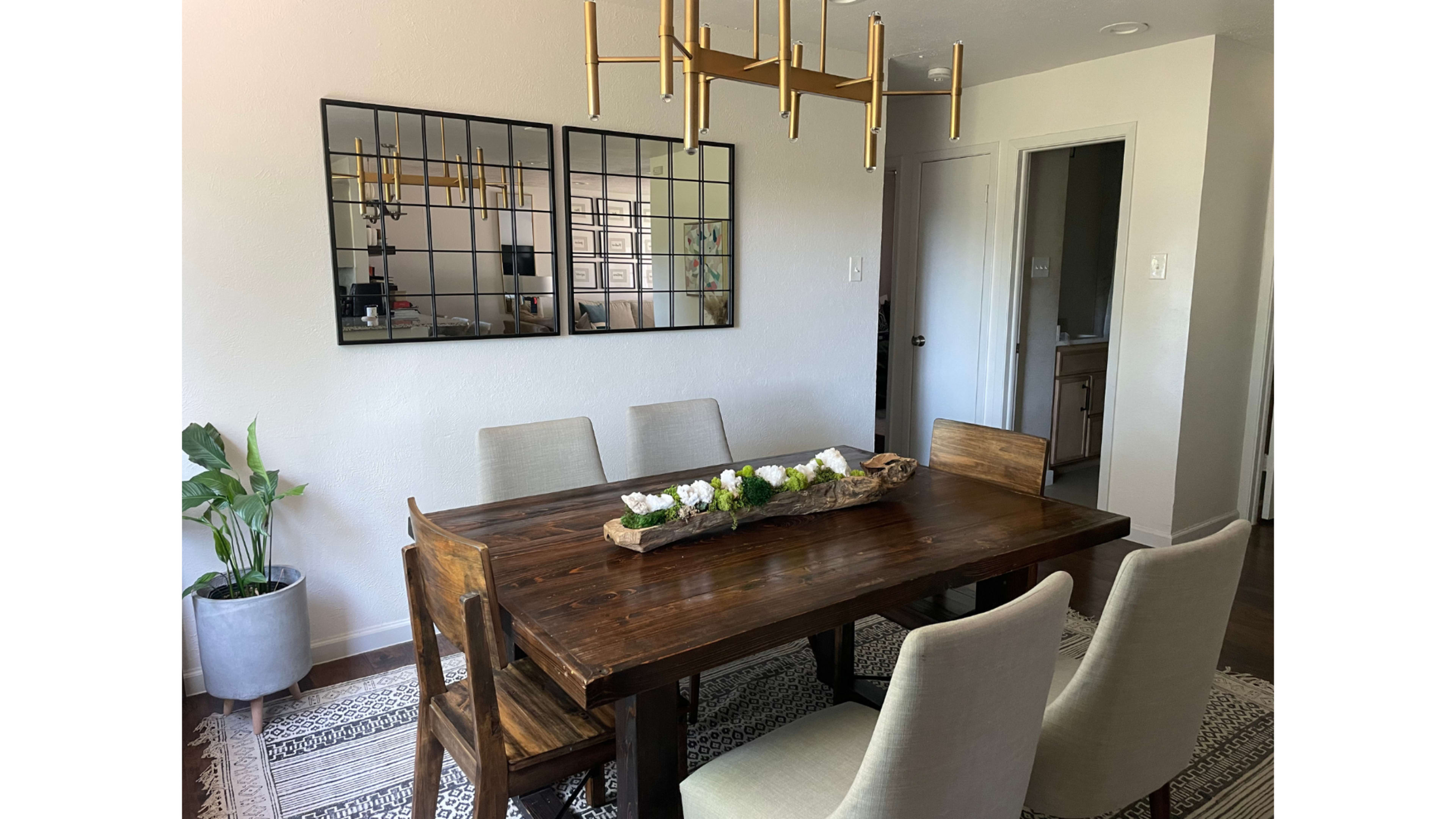 A wooden dining table with four upholstered chairs sits below a modern chandelier, accompanied by a decorative centerpiece and a large mirror on the wall.
