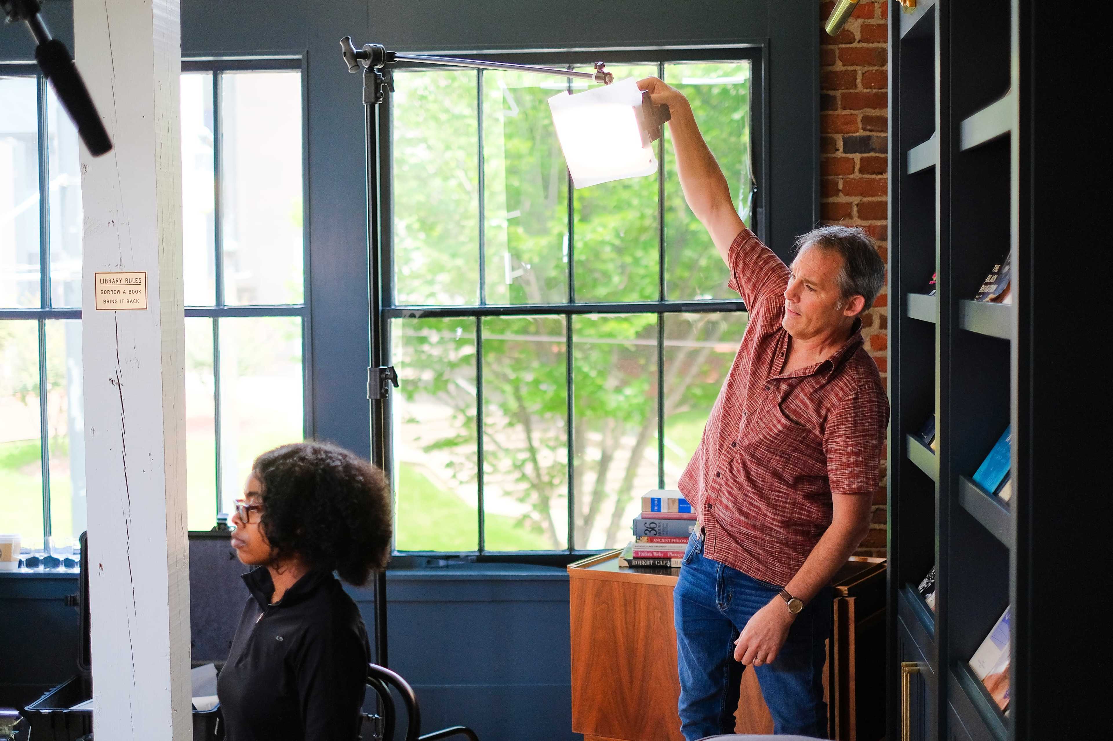 A man adjusts a light overhead while a woman sits in a chair nearby, both inside a room with large windows and bookshelves.