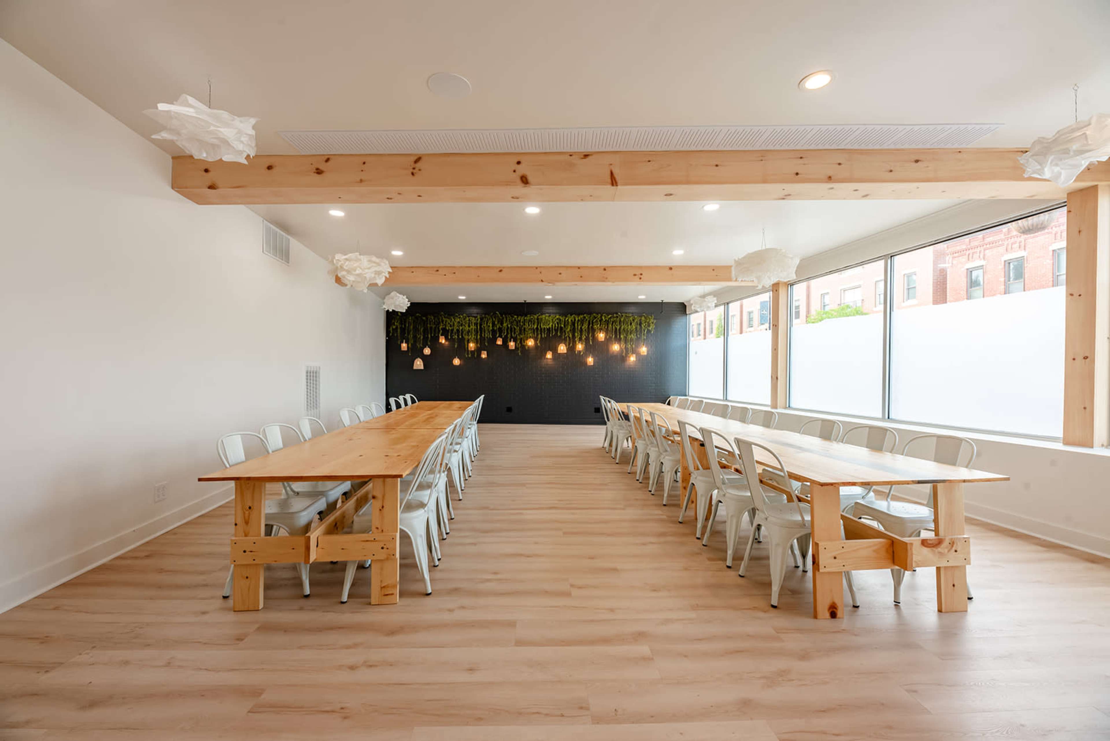 A spacious event room features long wooden tables arranged in rows, illuminated by hanging lights and adorned with greenery on a dark accent wall.