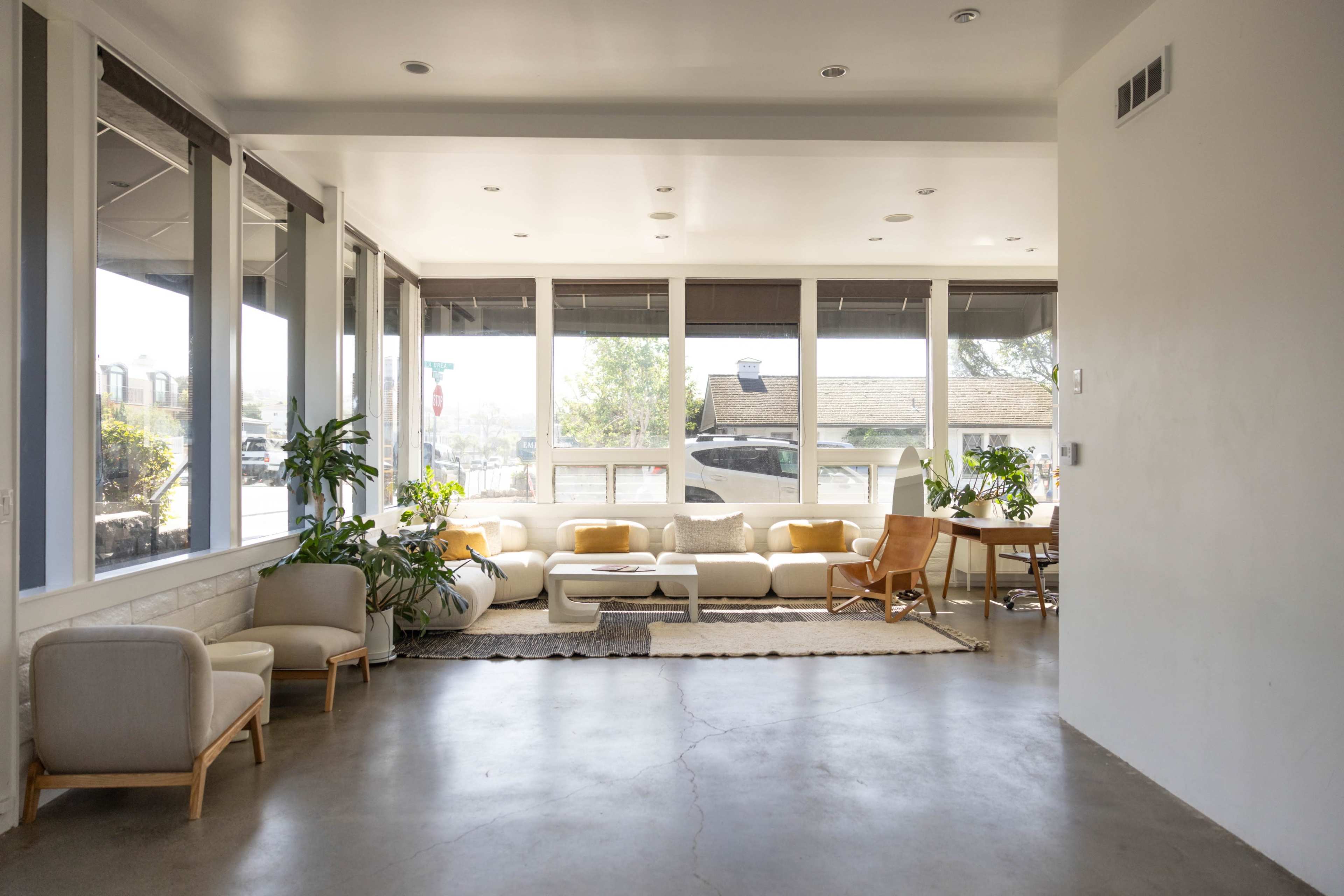 A bright, open living space features large windows, a light-colored sofa, minimalist furniture, and decorative plants.