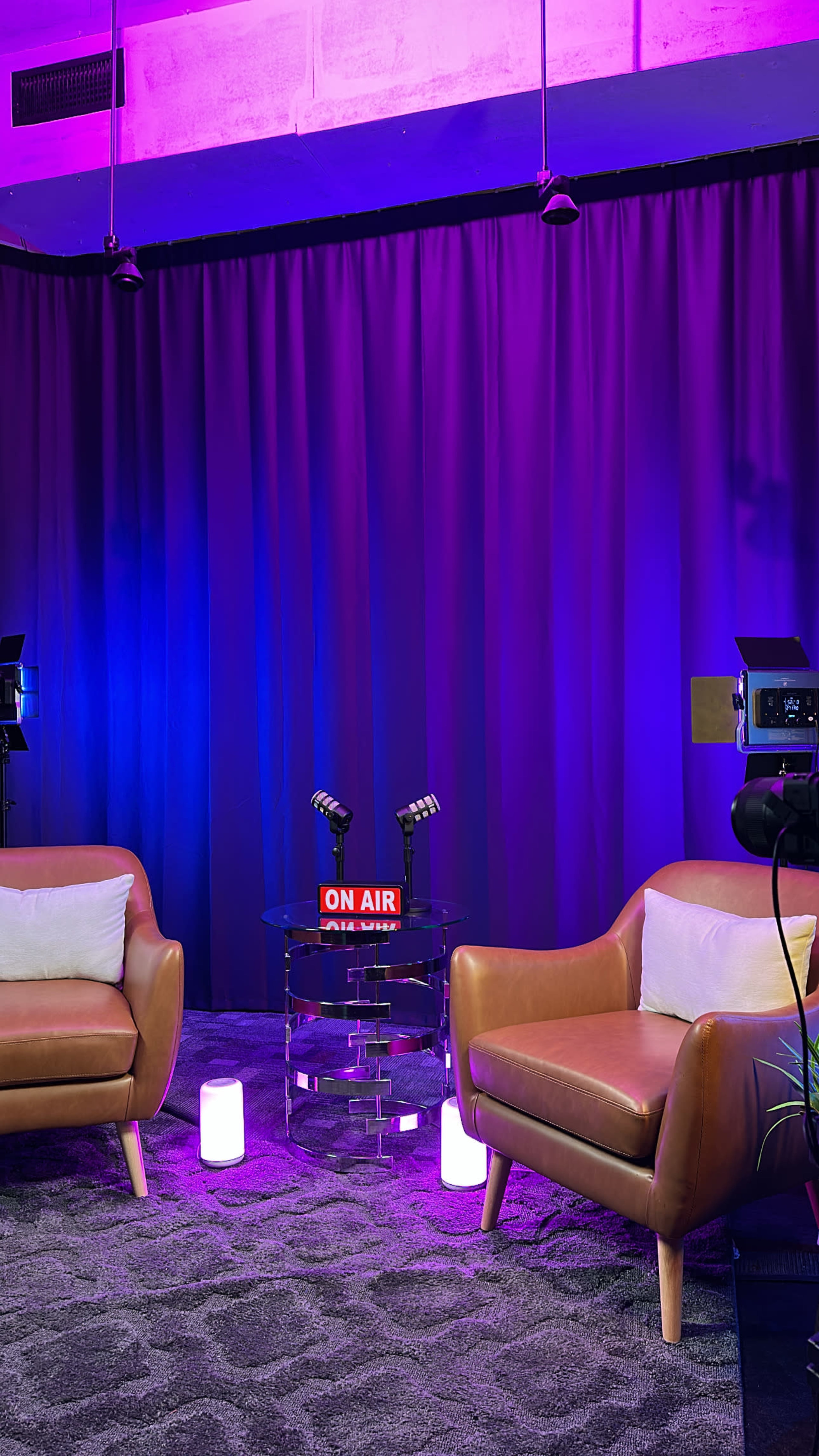The image shows a modern broadcasting or podcast studio with two brown armchairs, a clear glass table, and "ON AIR" signage against a backdrop of purple curtains and soft lighting.