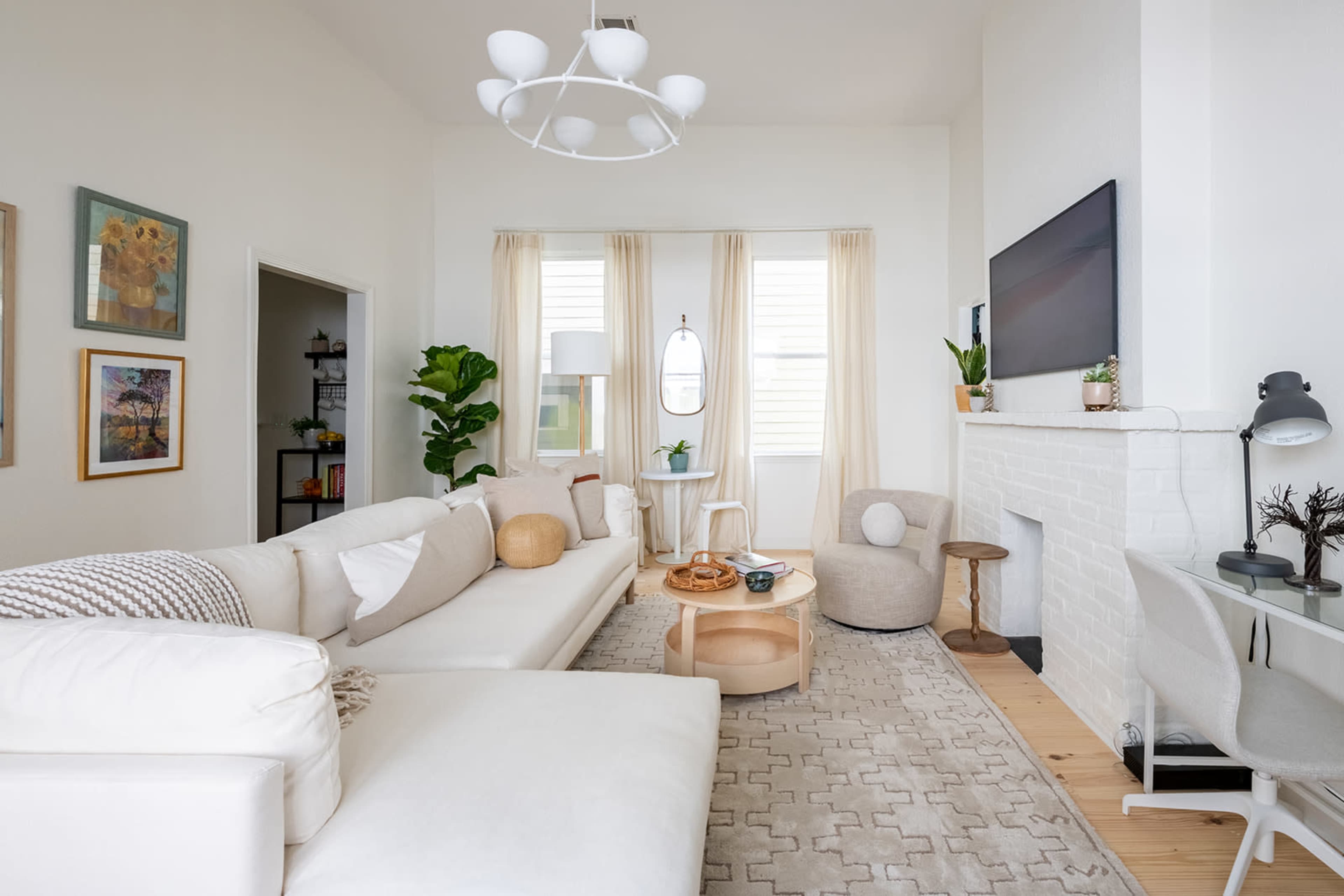 A spacious living room features a large white sofa, a light-colored rug, a round coffee table, and a modern light fixture, with large windows dressed in curtains.