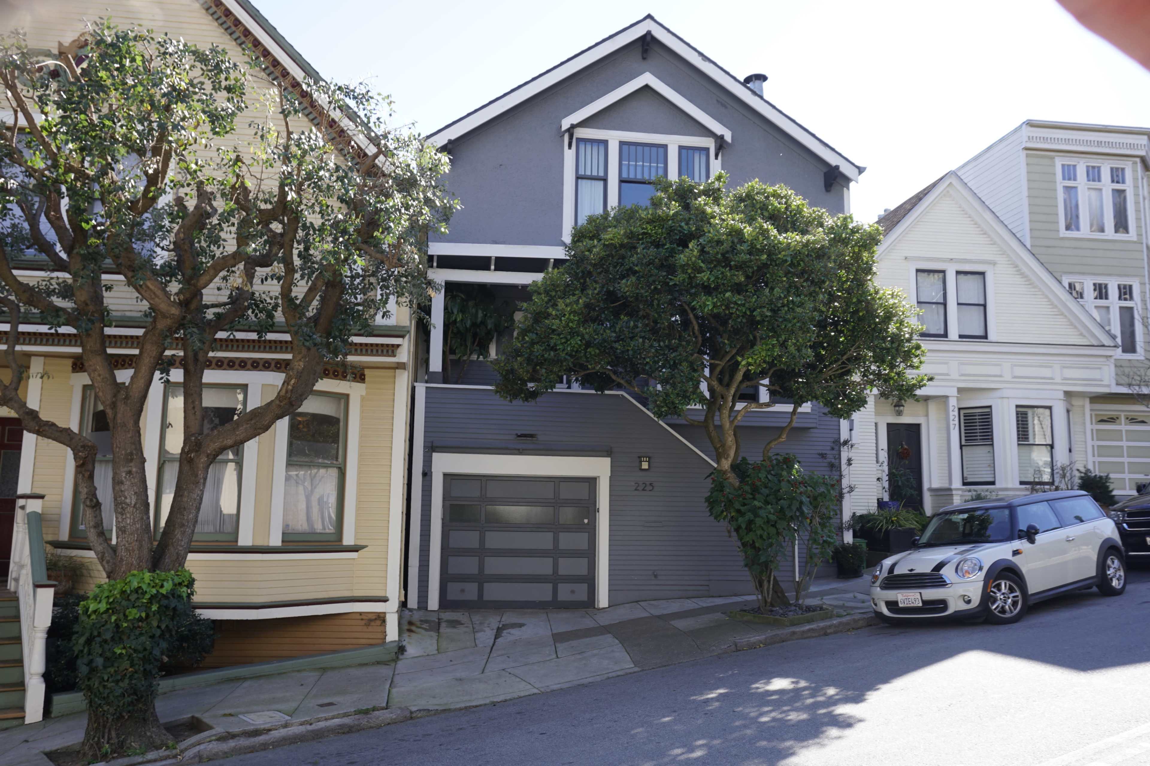 A residential street with several houses, featuring a gray house with a garage, flanked by trees and a parked mini car.