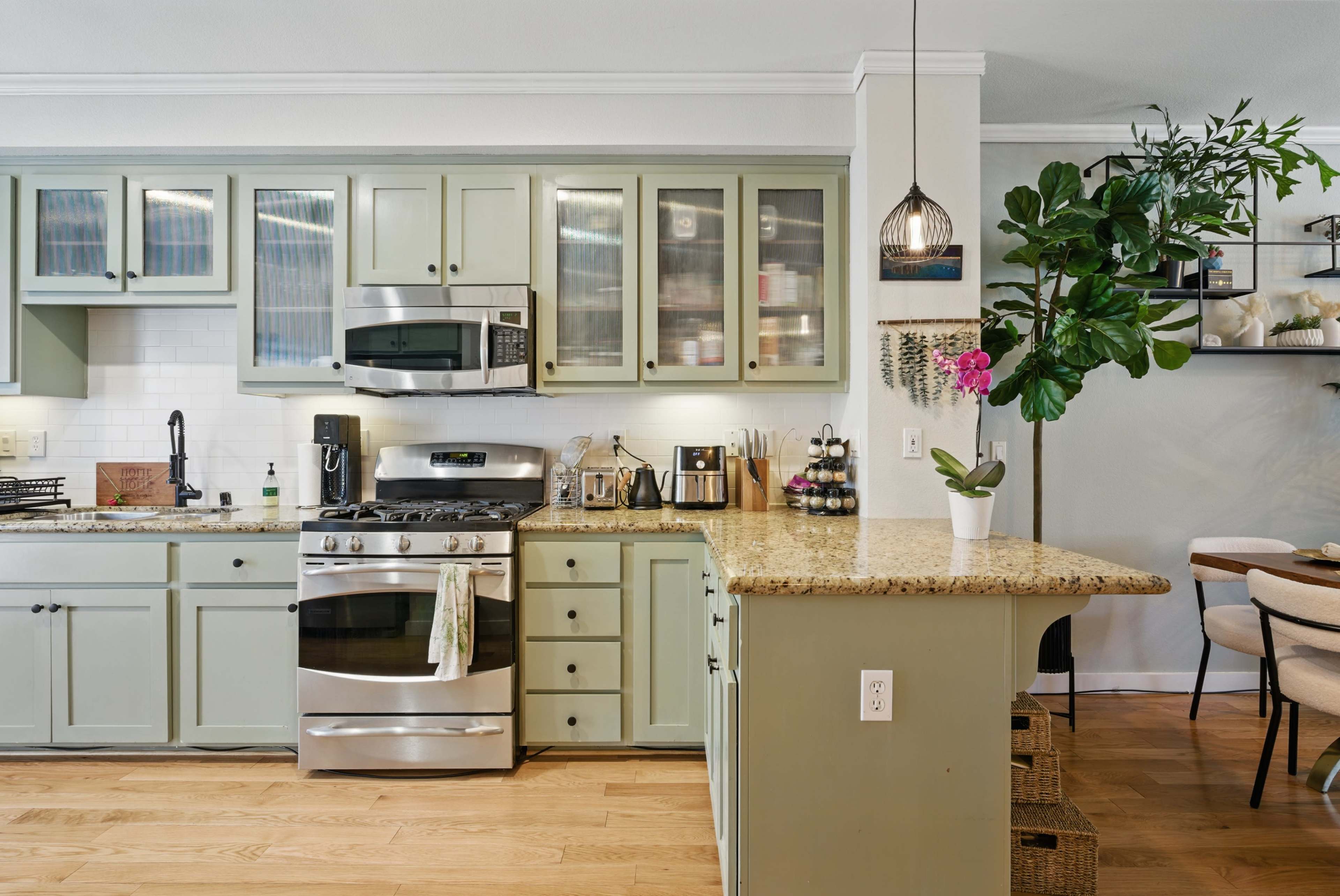 The image shows a modern kitchen with pale green cabinets, stainless steel appliances, and a granite countertop, featuring a dining area with plants and decor.