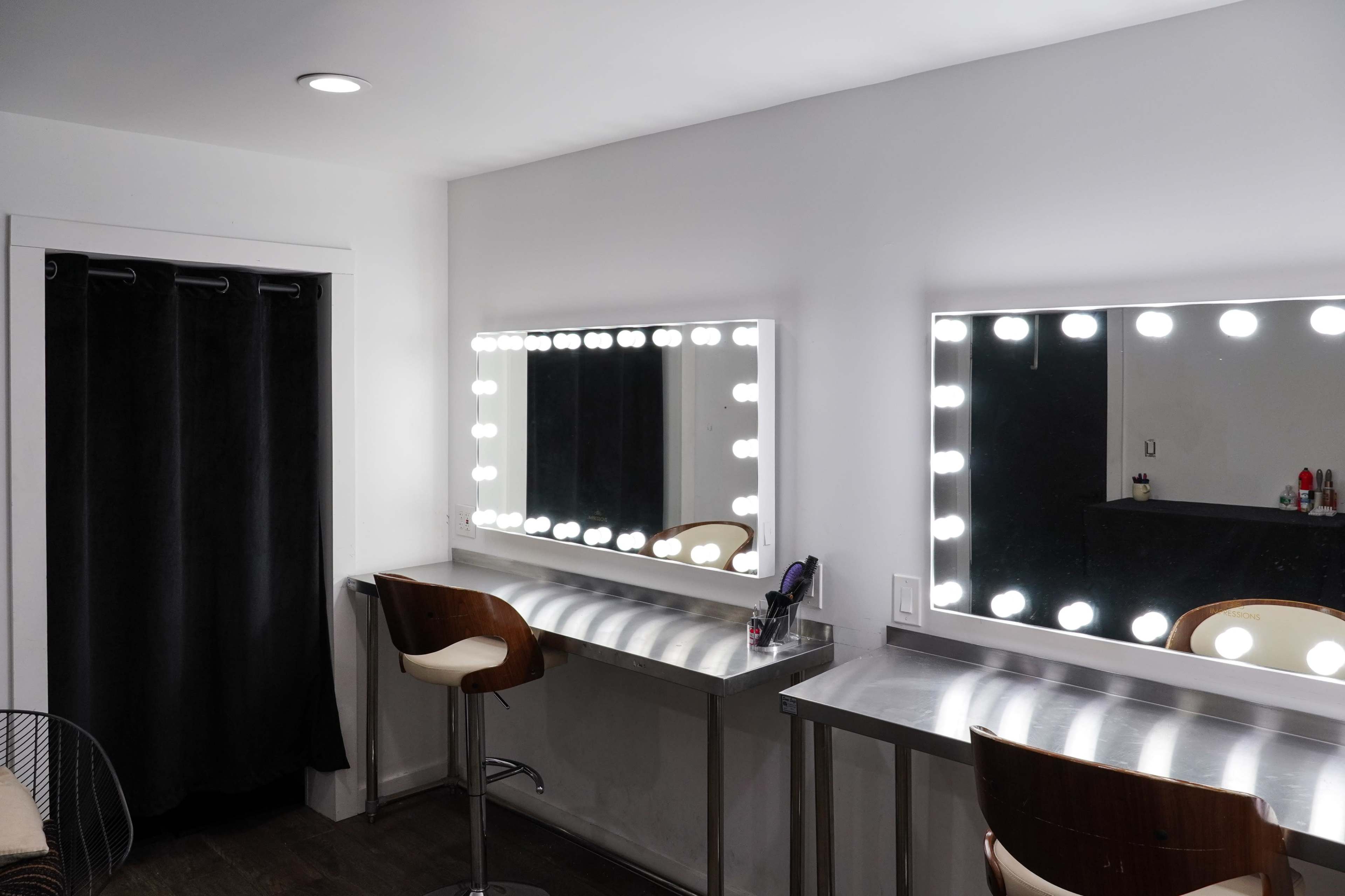 The image shows a well-lit makeup vanity area with two mirrors framed by lights, barstools, and a black curtain.