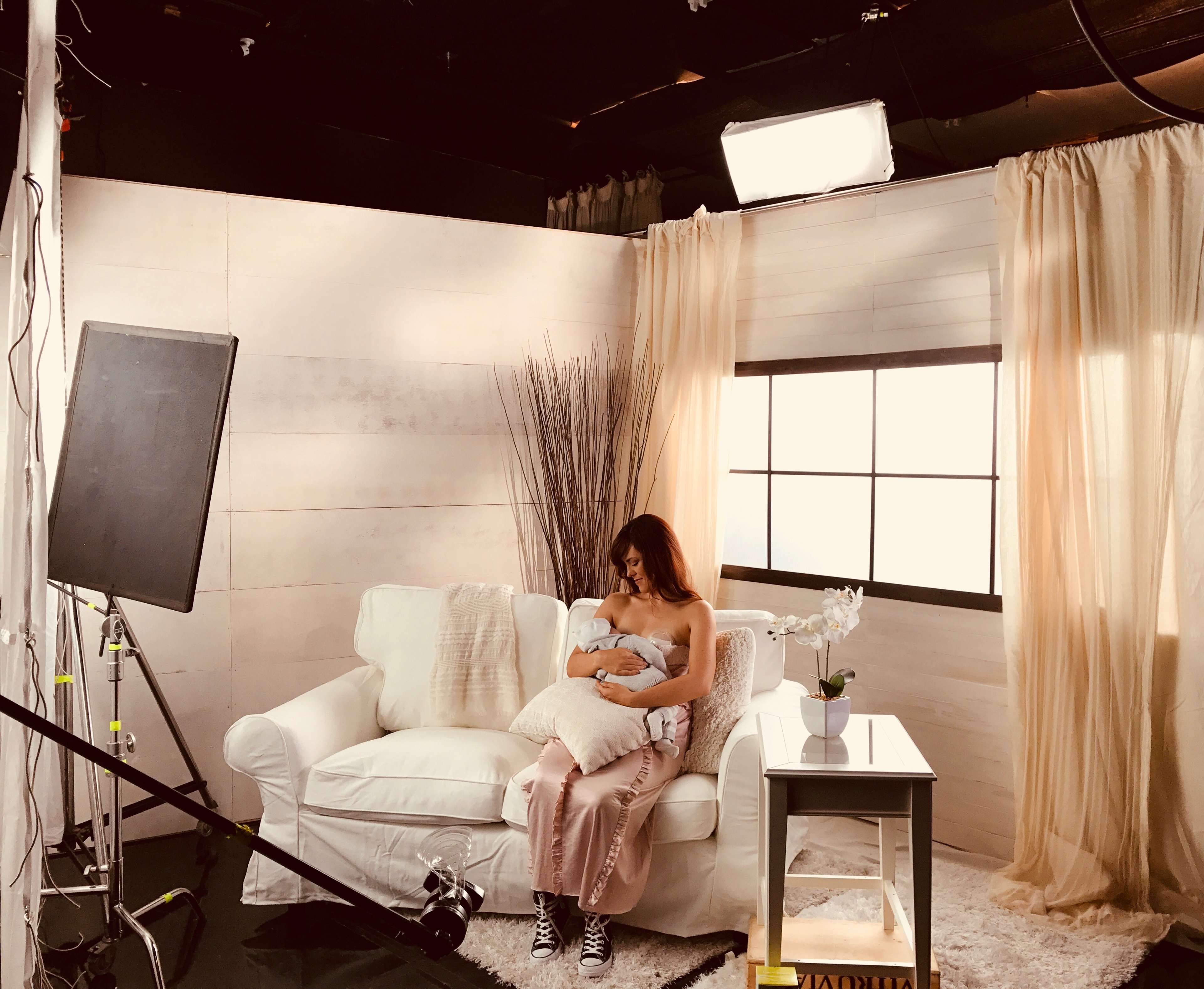 A woman sits on a white couch, holding a baby in a softly lit room with sheer curtains and a window in the background.