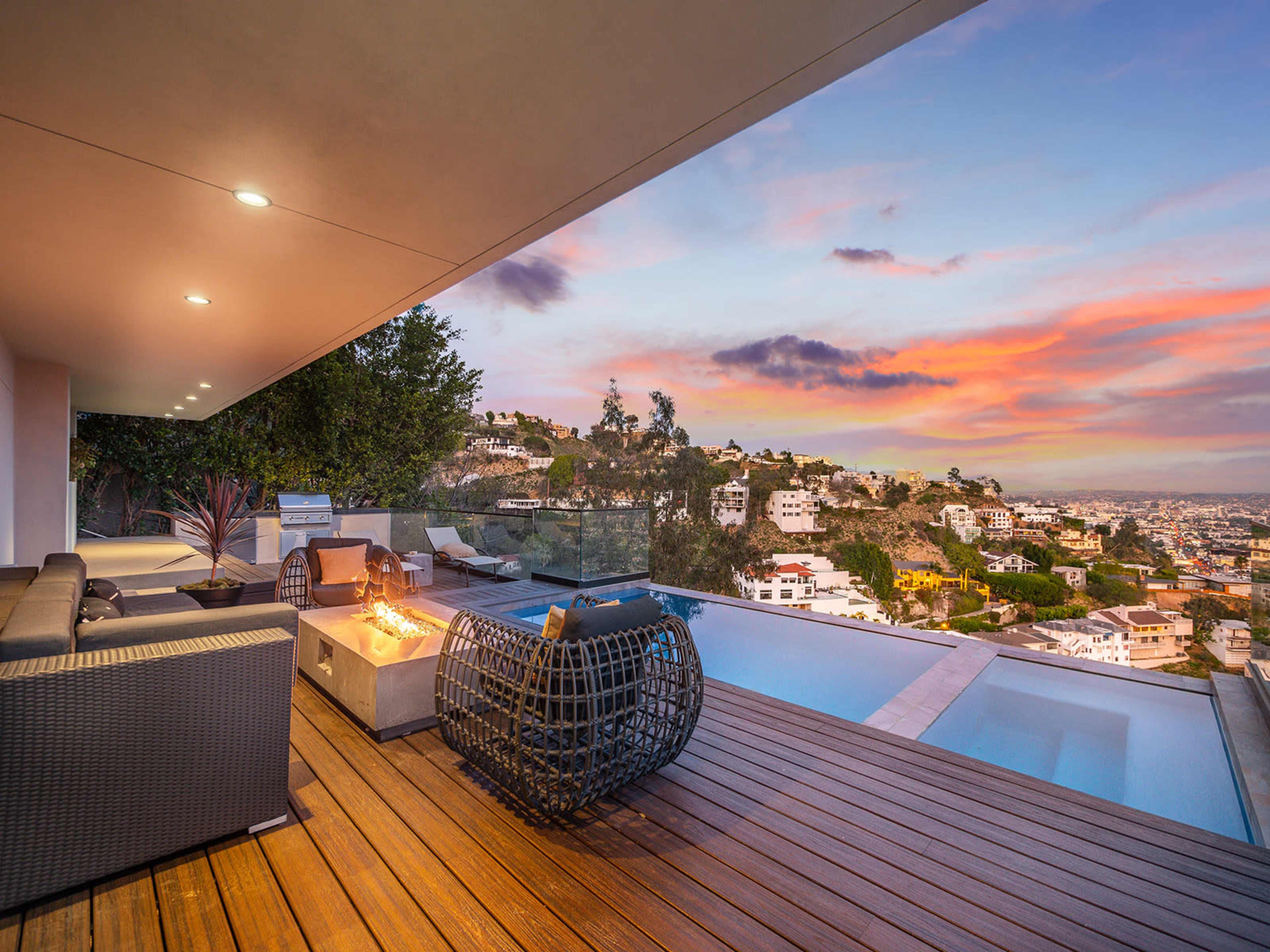 A modern outdoor lounge area features a fire pit and seating, overlooking a hillside with houses at sunset.