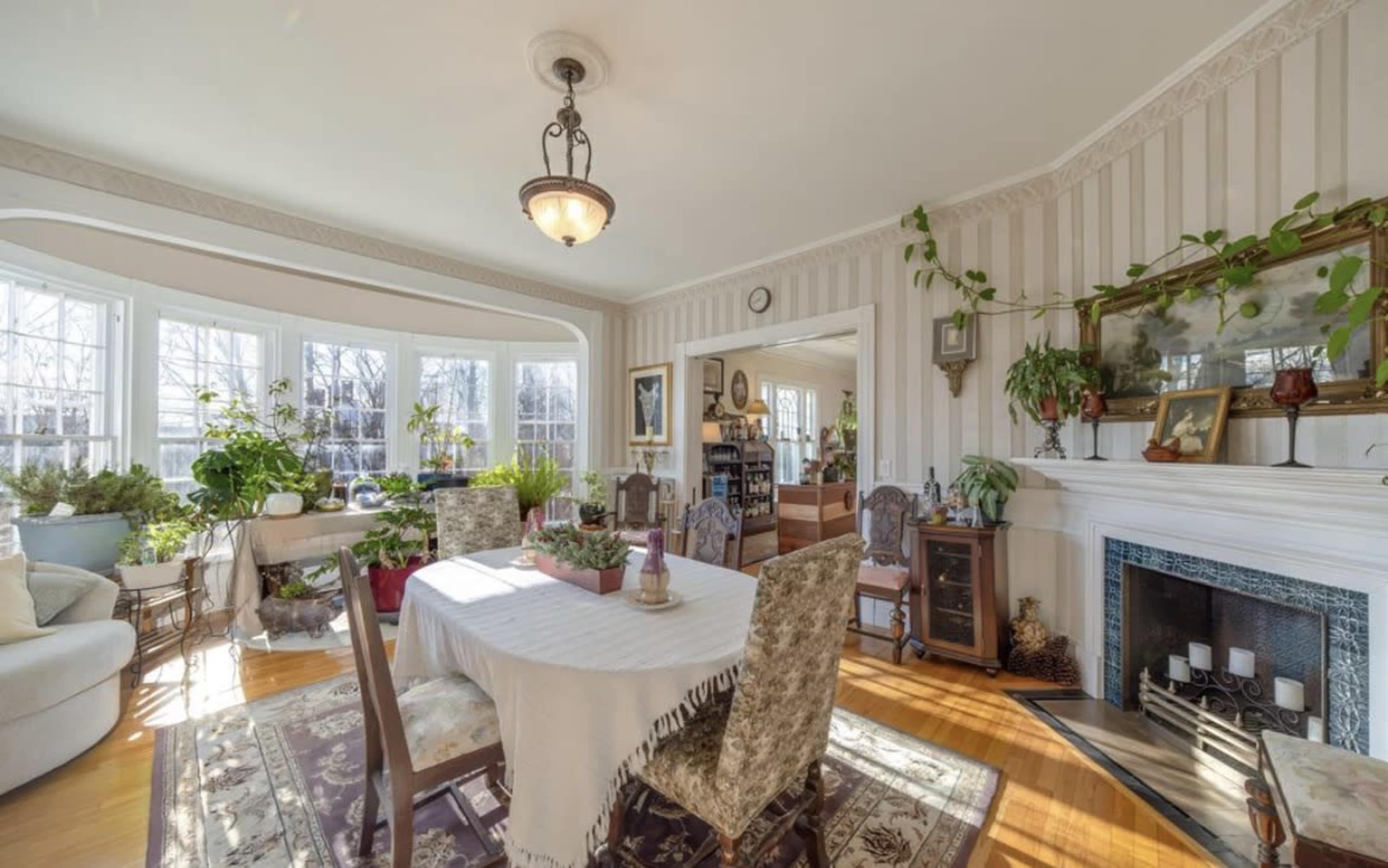 The image features a bright dining room with a large table surrounded by chairs, a fireplace, and multiple plants near large windows.