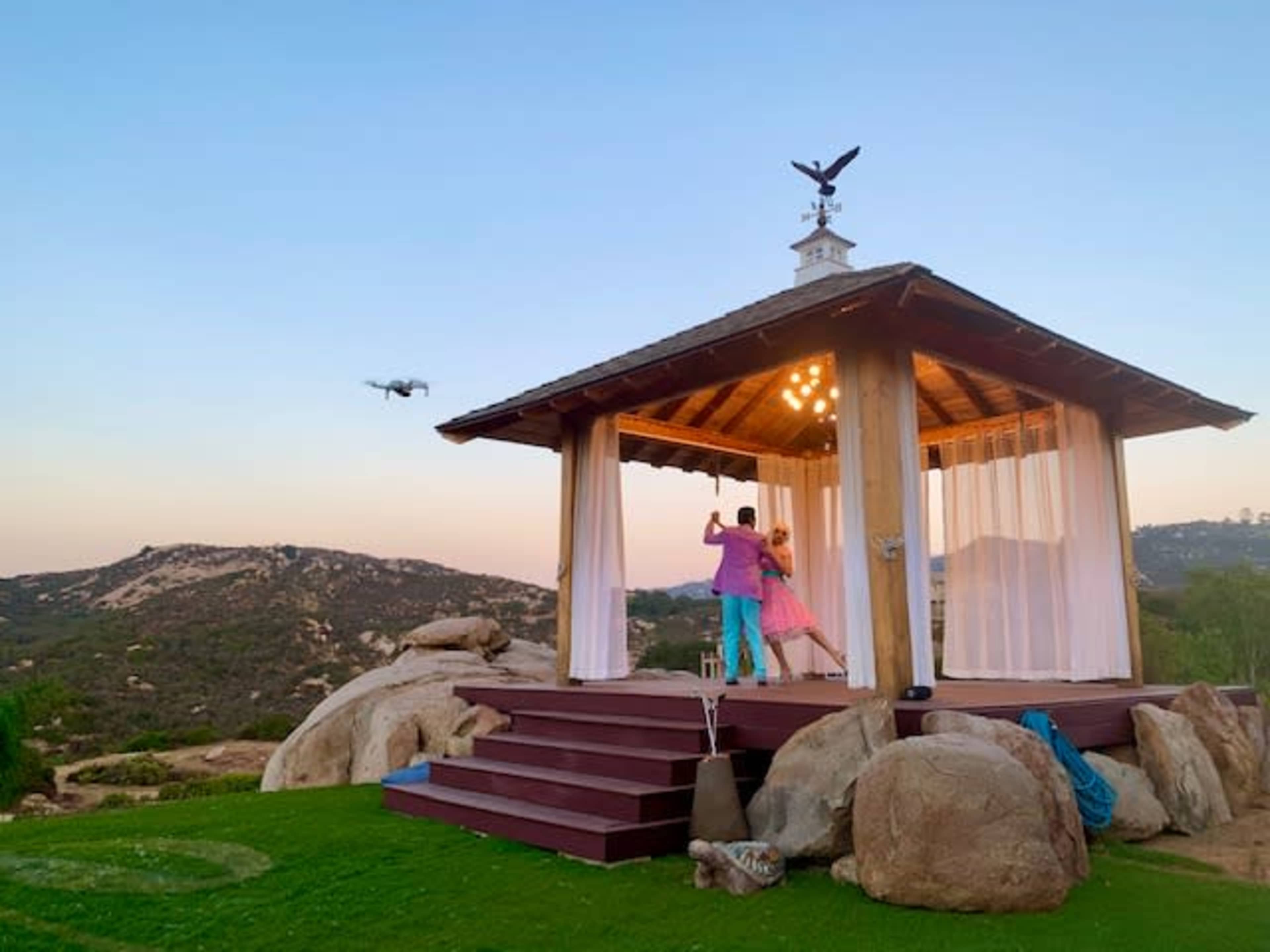 A couple dances under the pavilion at sunset, with a drone flying above in a mountainous landscape.