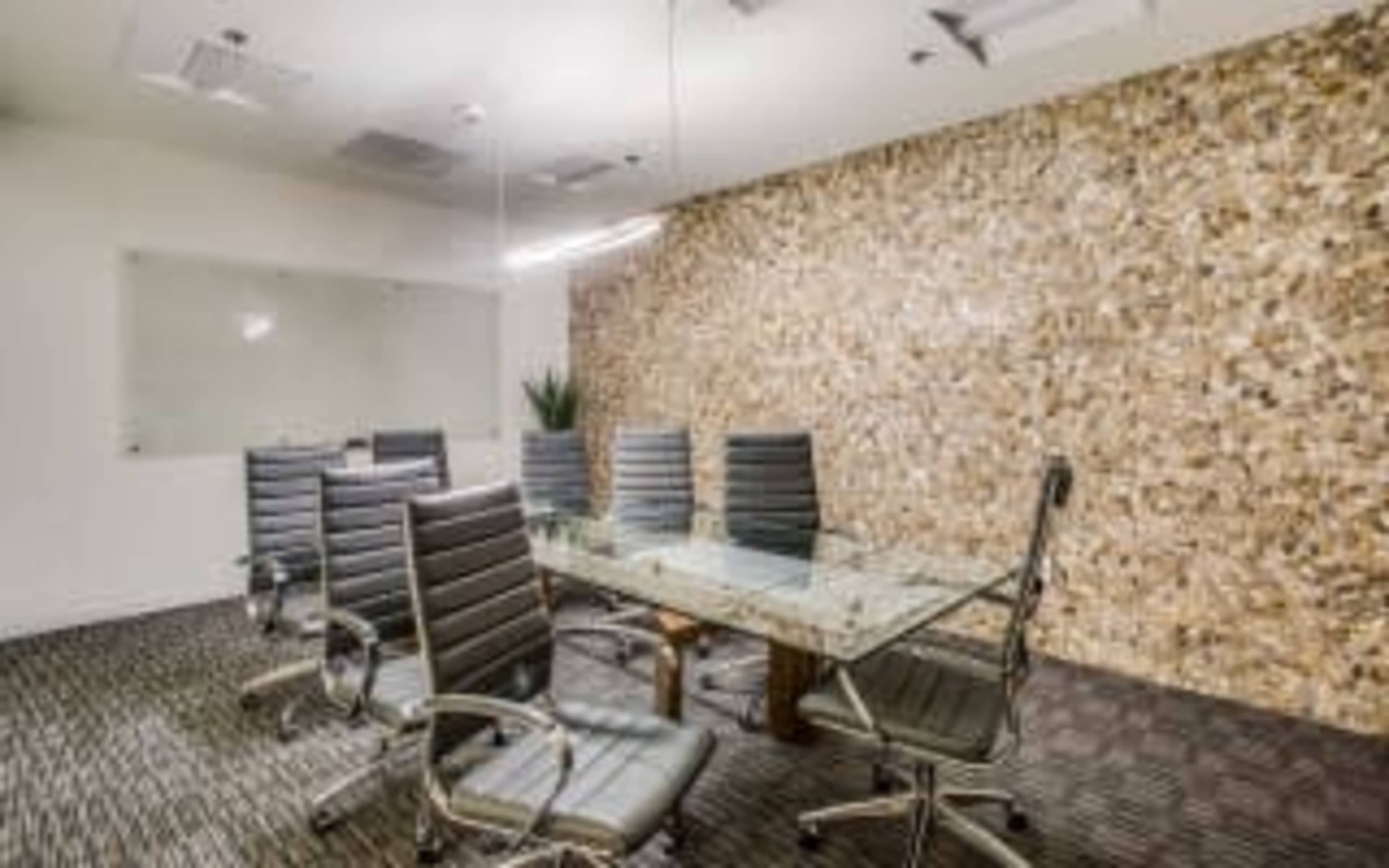 The image shows a modern conference room with a glass table, six leather office chairs, and a textured stone wall.