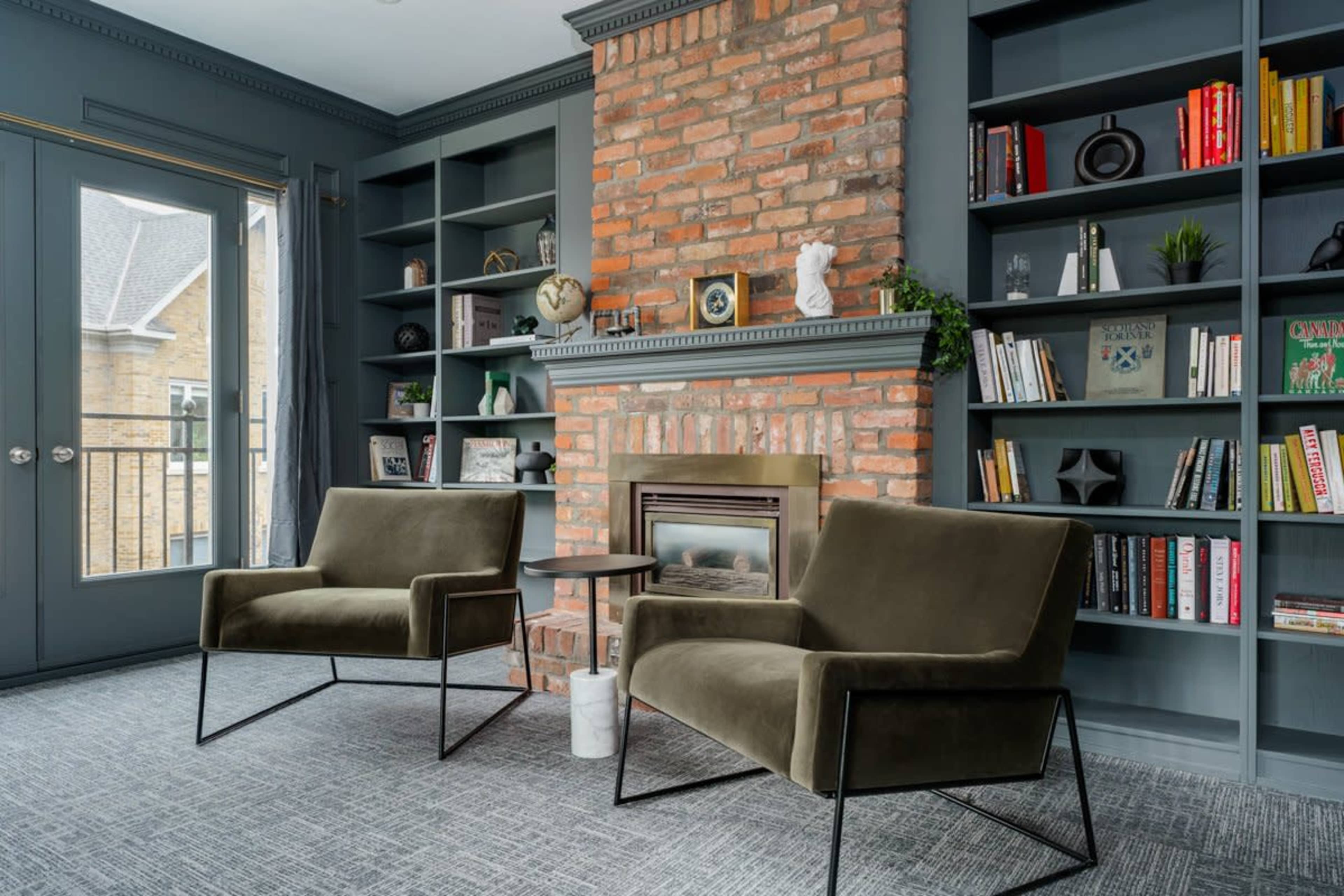 A cozy reading nook features two green chairs, a small table, and a brick fireplace surrounded by built-in bookshelves filled with books and decorative items.