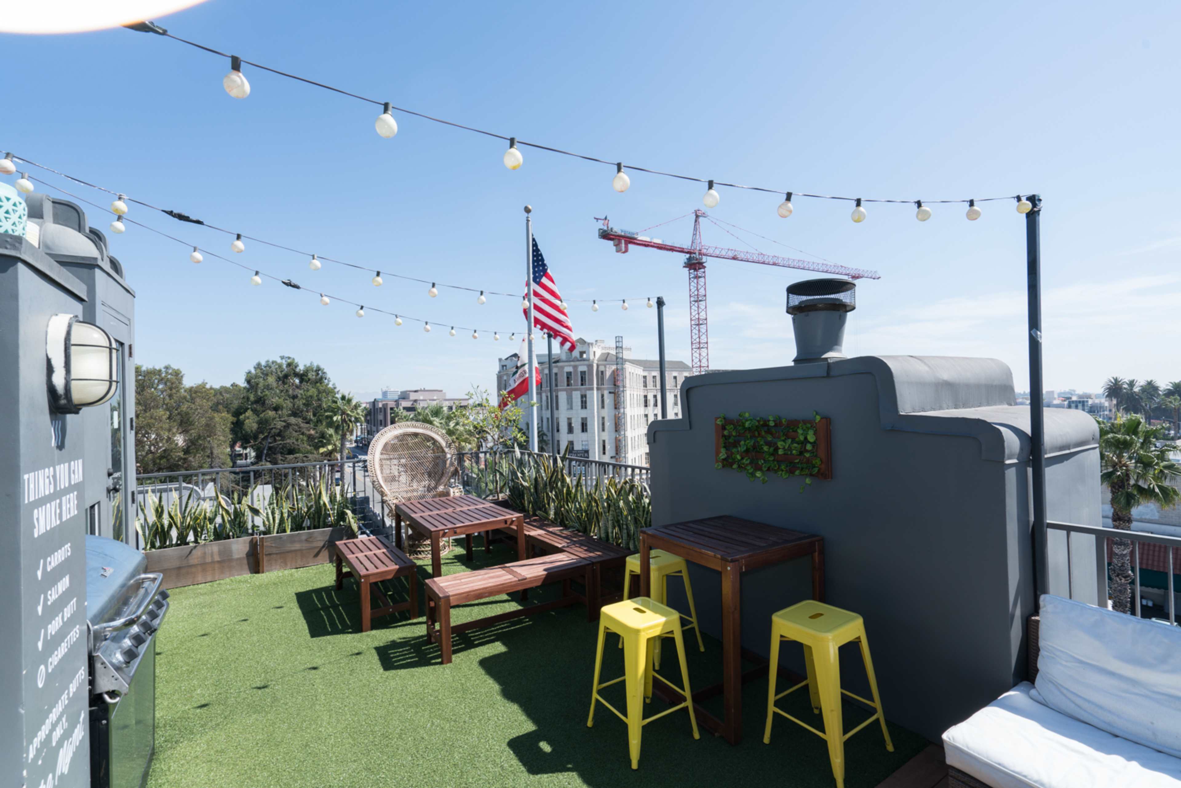 A rooftop terrace features wooden seating, yellow stools, and string lights, with an American flag in the background and a construction crane visible nearby.