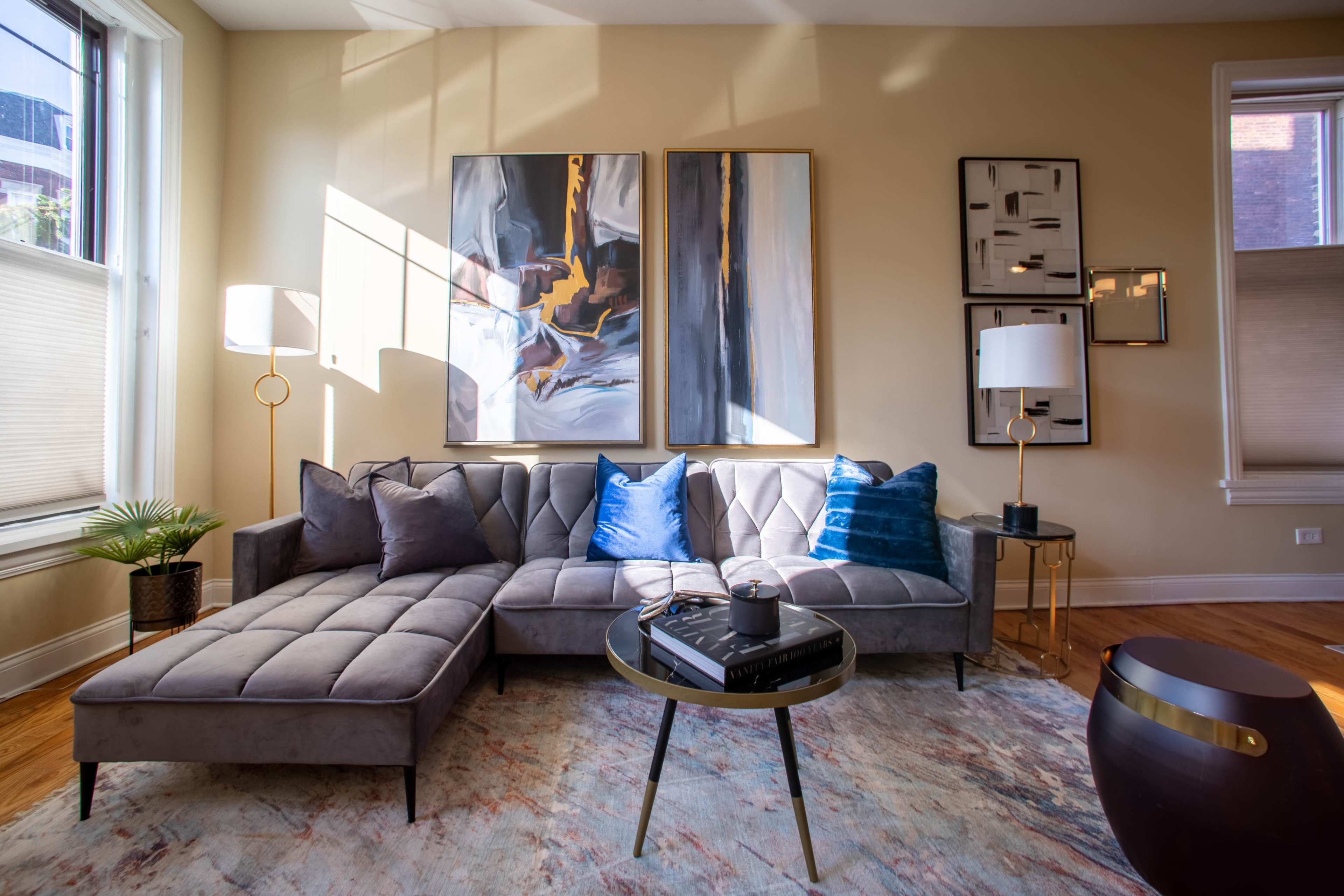 A modern living room features a gray sectional sofa, two abstract paintings on the wall, a round coffee table, and a floor lamp, with natural light streaming through the windows.