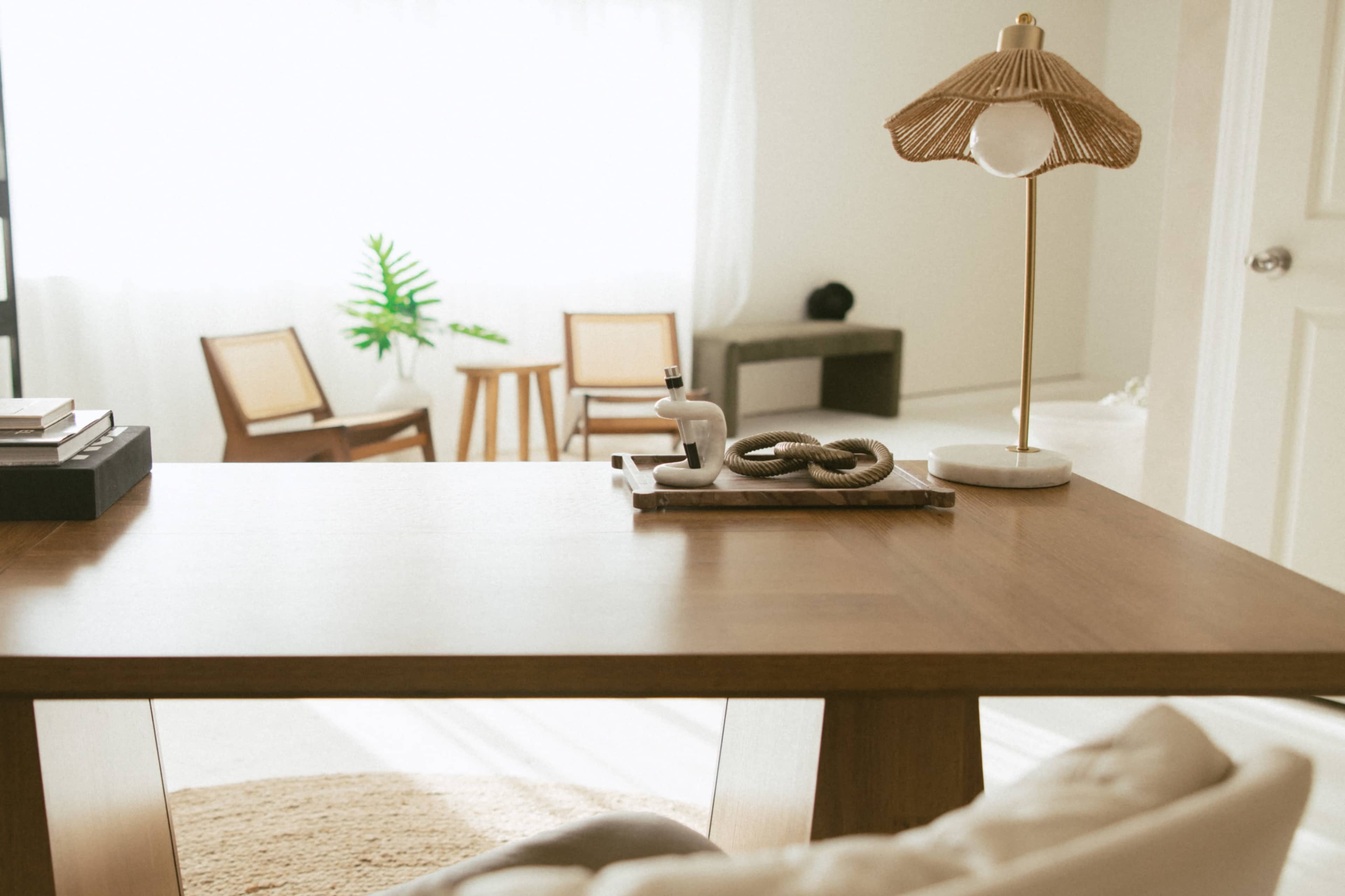 A wooden desk with a lamp and decorative items is positioned in a bright room featuring minimalistic chairs and a plant in the background.