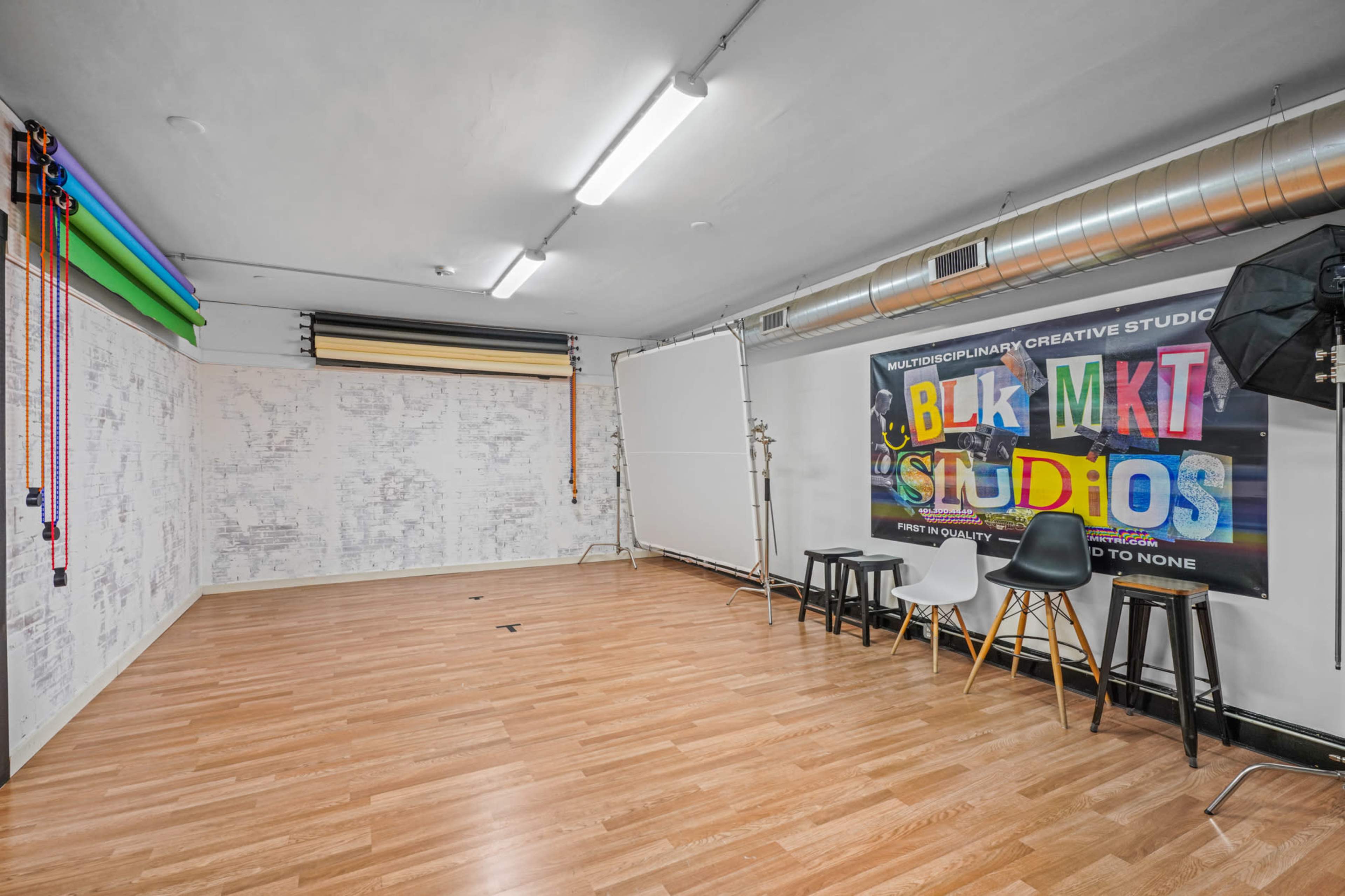 A spacious studio with wooden flooring, bright overhead lighting, and a large colorful banner featuring the text "BLK MKT STUDIOS" mounted on the wall.