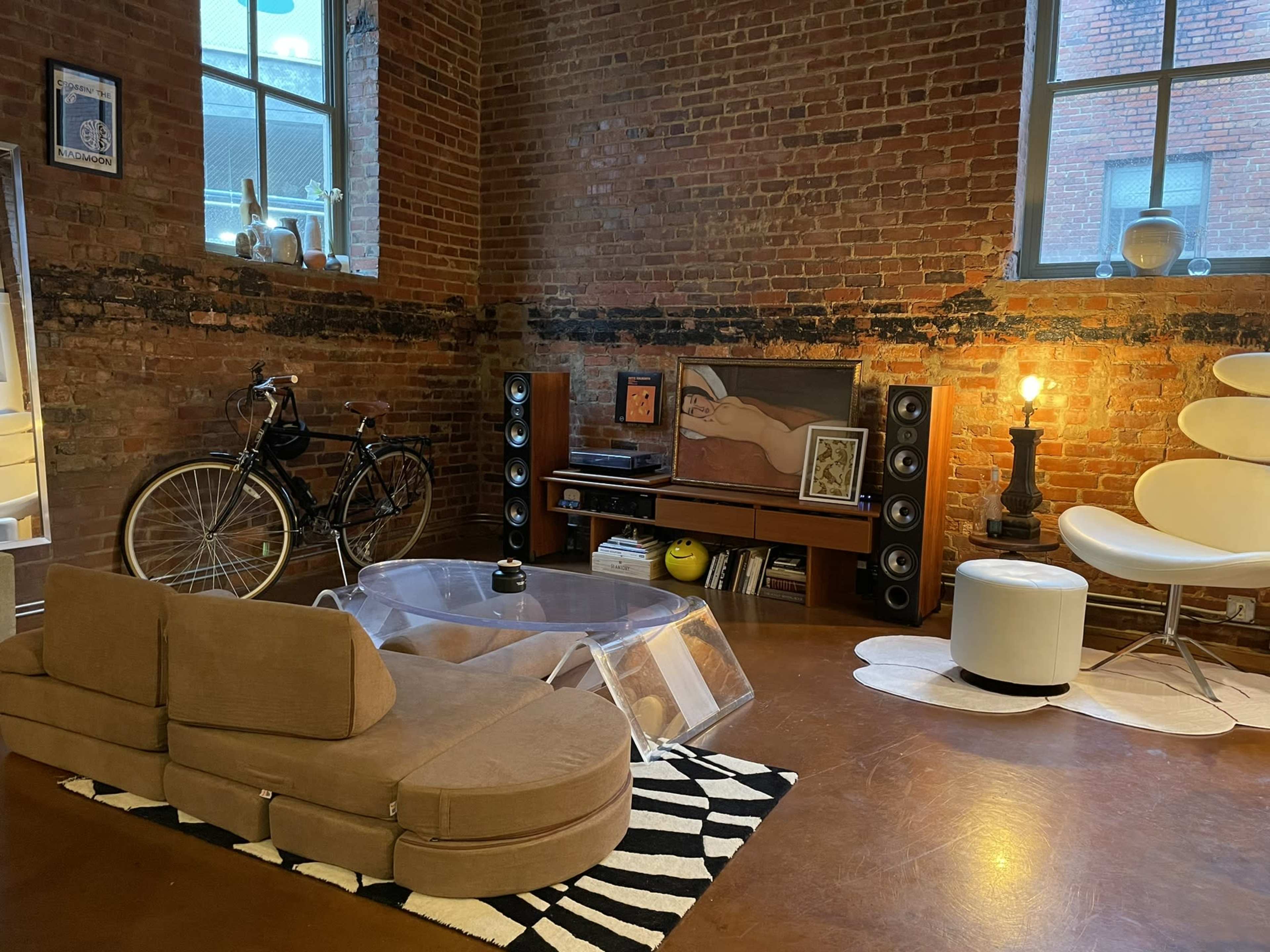 A cozy living space features a bicycle, a curved sofa, a clear coffee table, a media unit with speakers, and an iconic chair beside a lamp against a brick wall.
