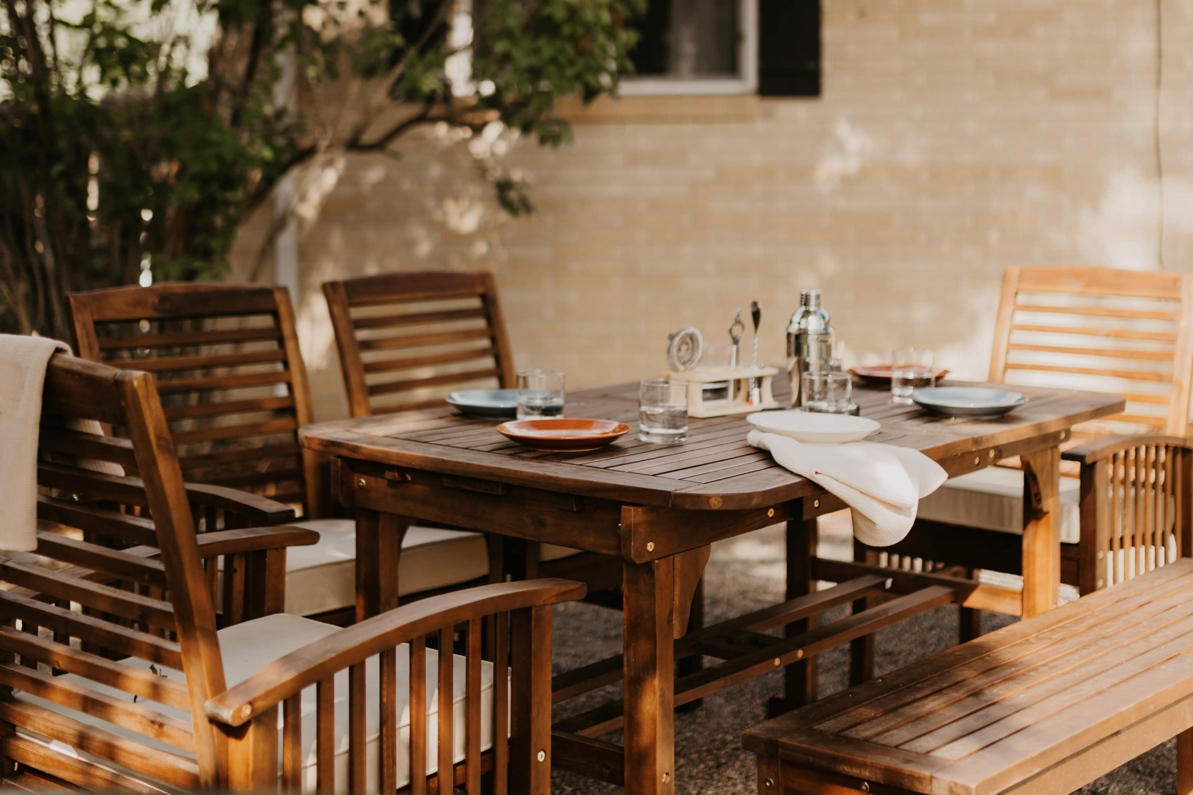 A wooden dining table is set outdoors, surrounded by chairs, with glassware and plates arranged for a meal.