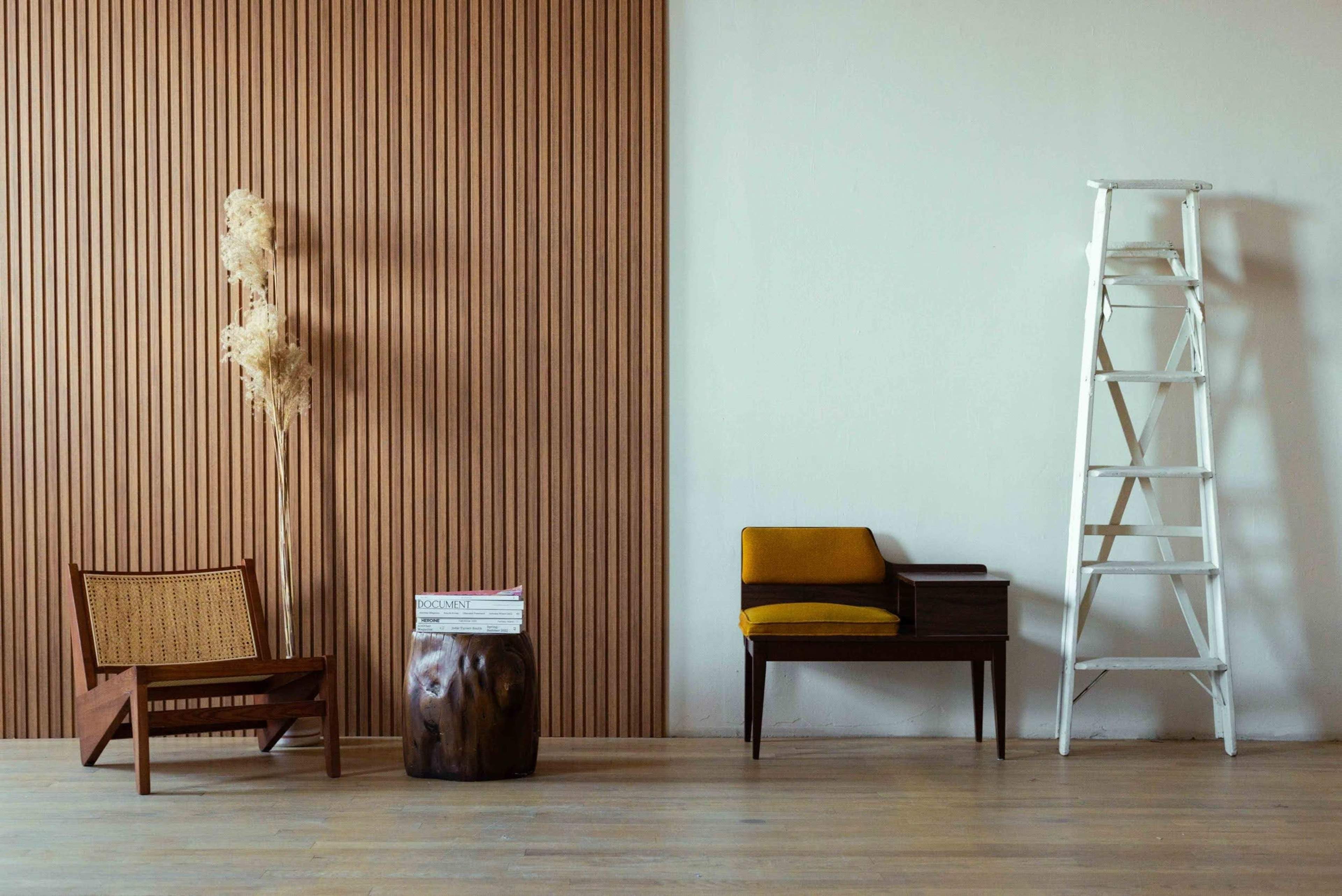The image shows a minimalist interior with a wooden wall feature, a small ladder, a brown accent stool, and a wooden chair with yellow upholstery.