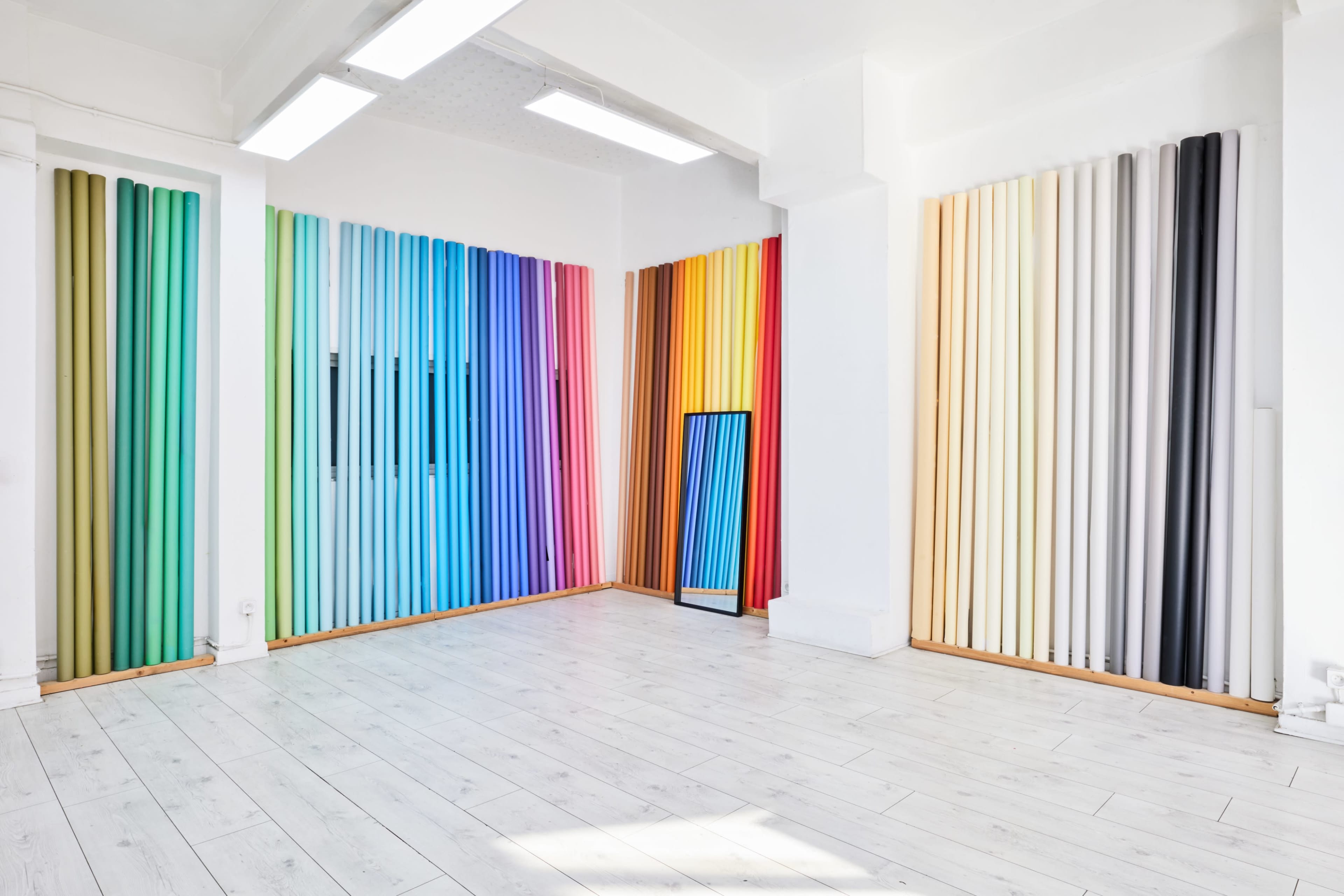 The image shows a bright, spacious room with a white floor and walls, featuring vertical strips of colorful paper or fabric arranged on the walls and a mirror in one corner.