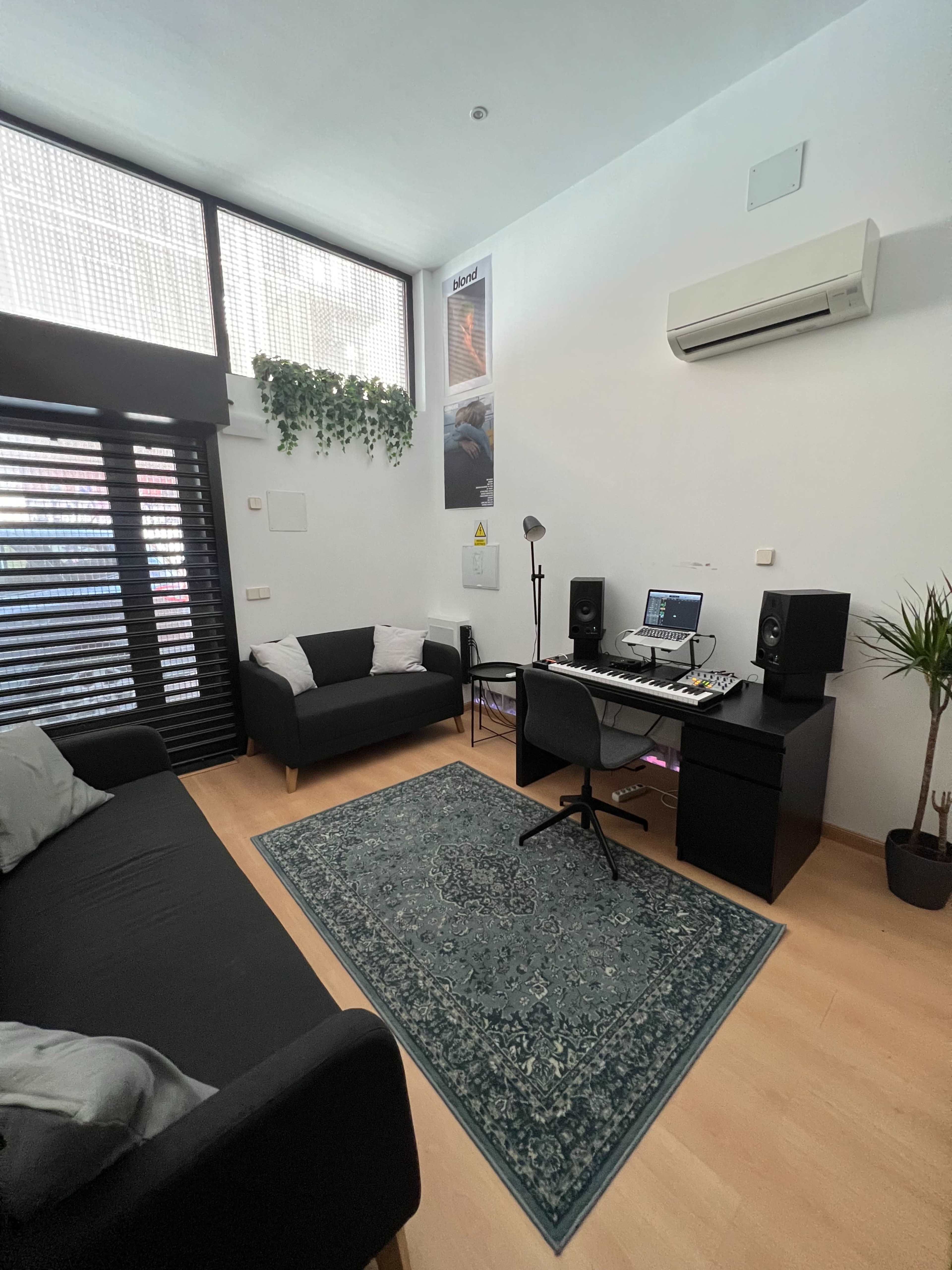 A small room with a desk featuring audio equipment, two sofas, a rug, and a plant, illuminated by natural light from a large window.