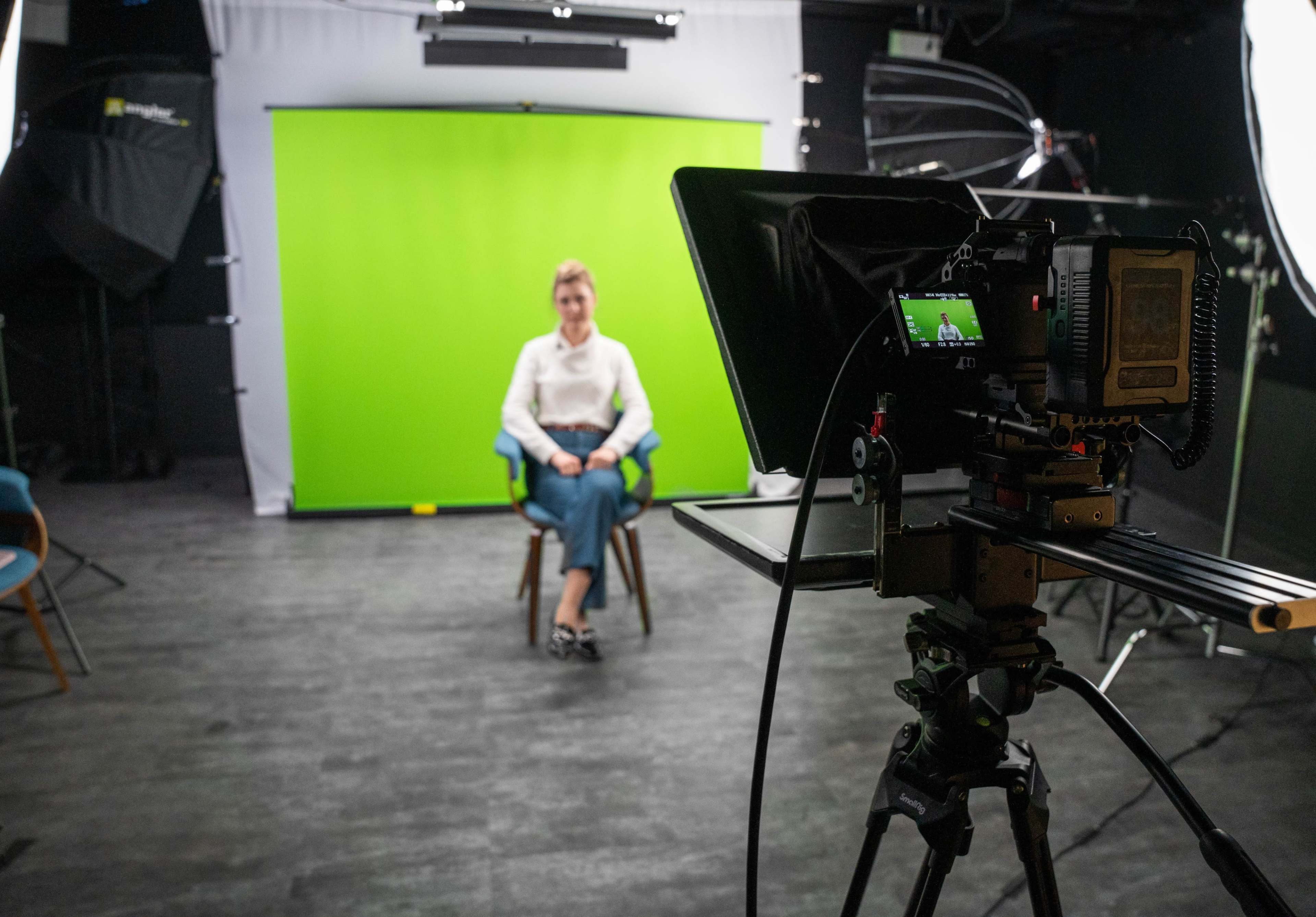 A woman sits in front of a green screen in a photo studio, while a camera on a tripod is focused on her.