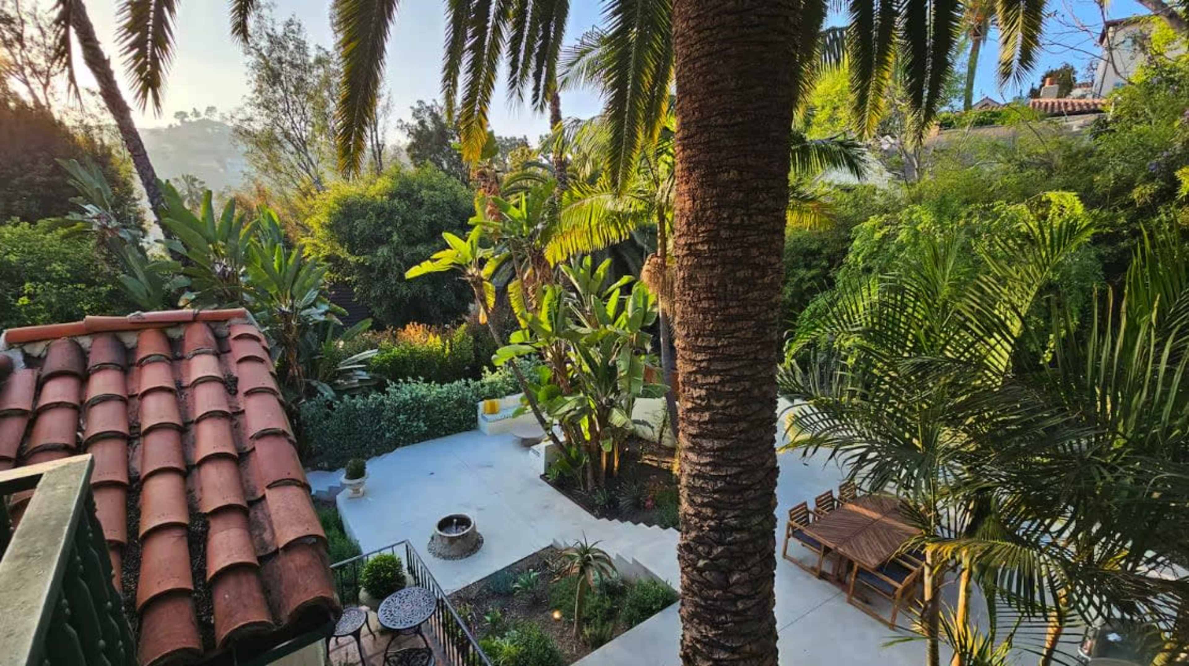 The image shows a landscaped garden with various tropical plants, a seating area, and a stone patio, viewed from an elevated position.