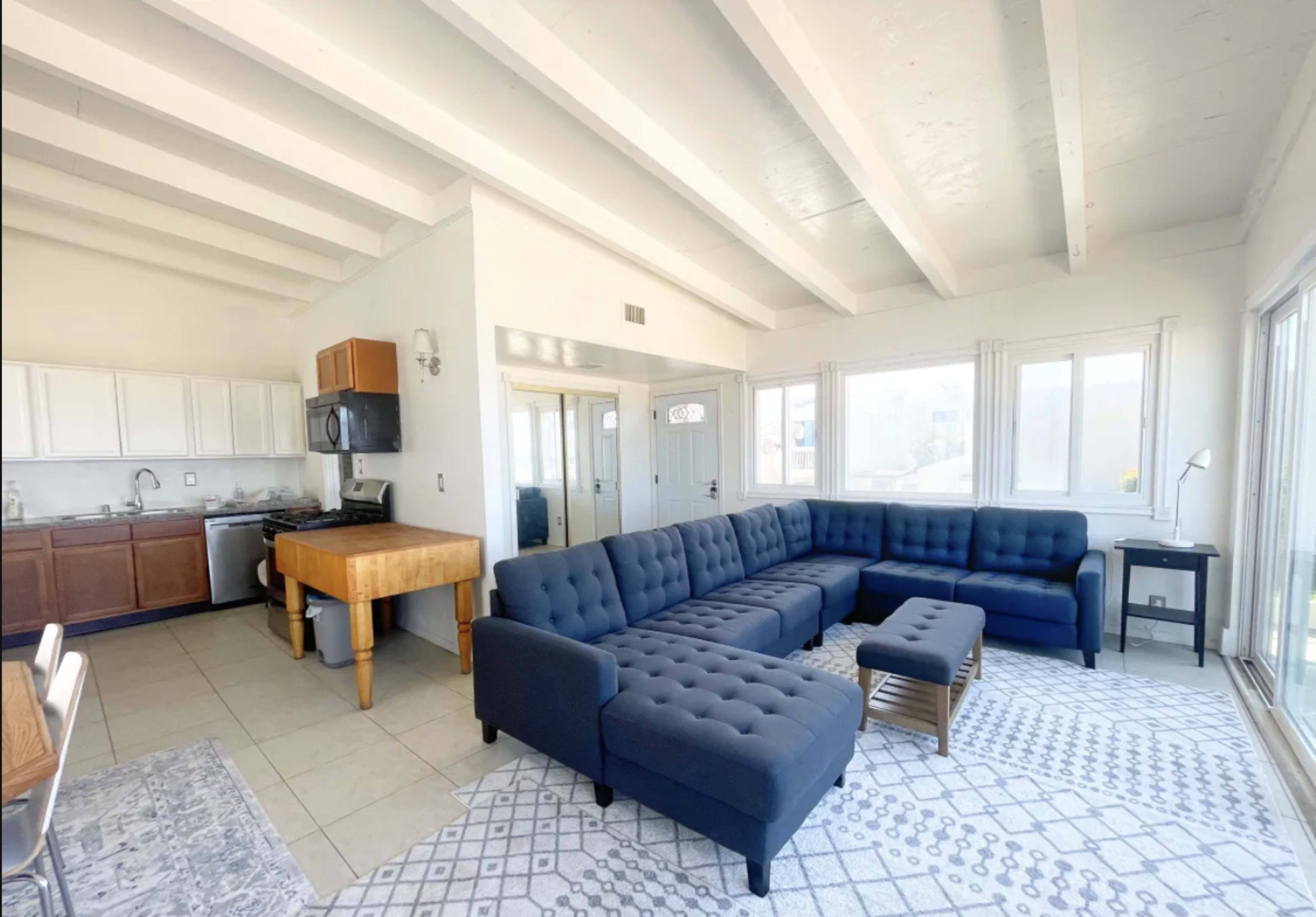 A modern living room with a dark blue sectional sofa, a wooden table, and a kitchen area in the background.
