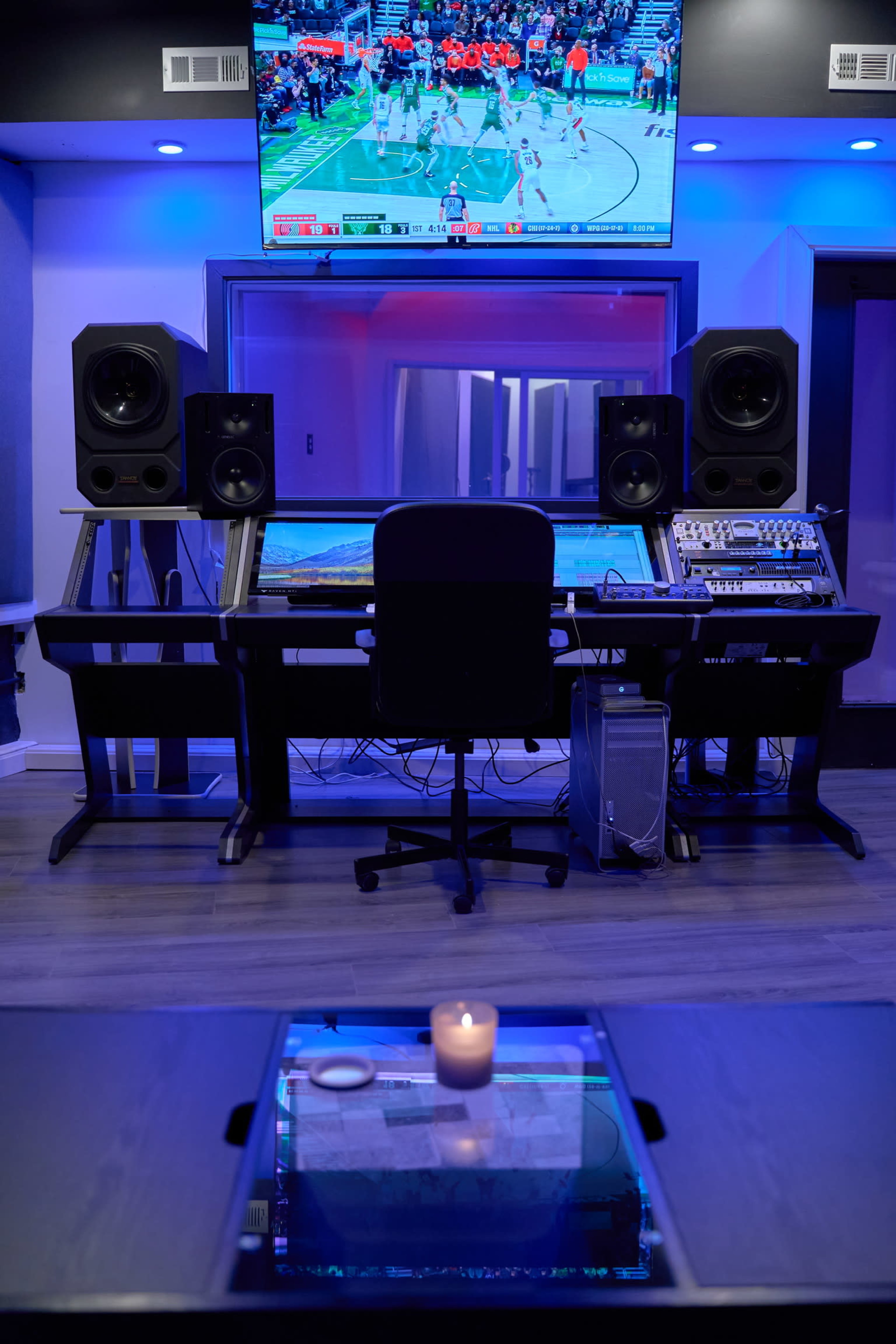 A modern recording studio features a sound mixing console with large speakers, a television displaying a basketball game, and a table with a candle in front.