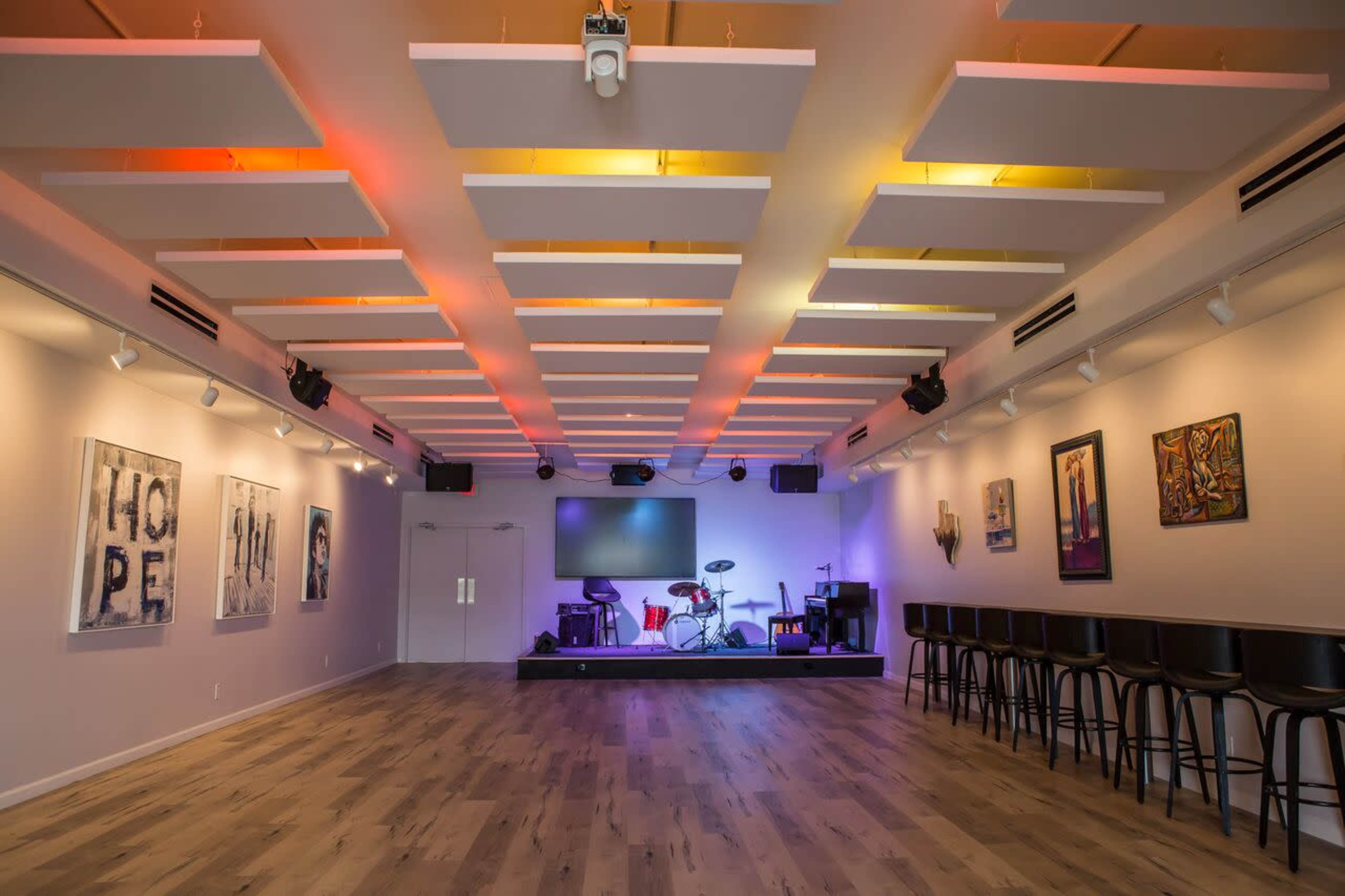 The image shows a spacious, modern performance venue with a stage, drum set, and artwork on the walls.