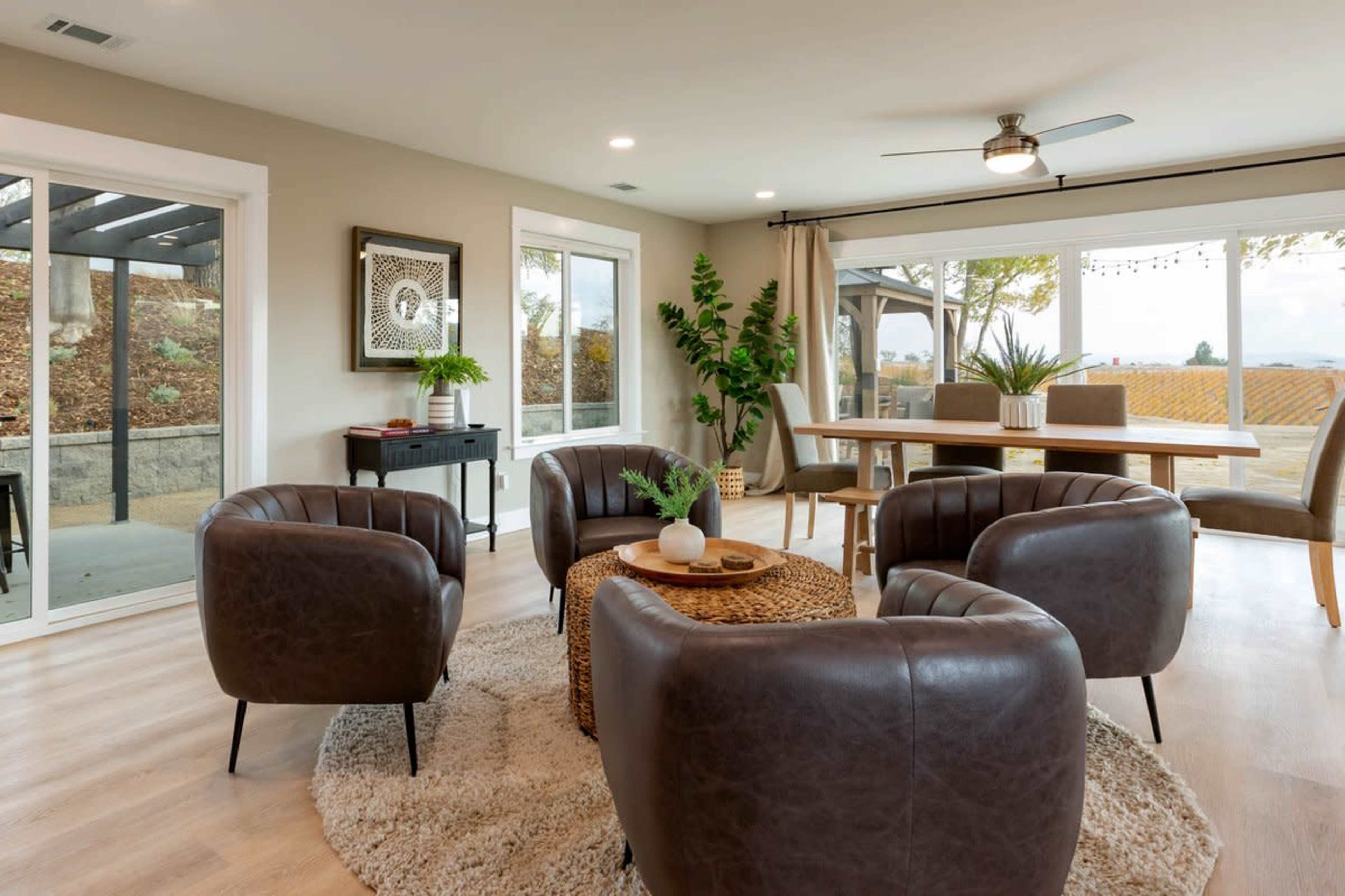 A modern living room with four brown leather chairs arranged around a circular coffee table on a light rug, with large windows overlooking a vineyard.