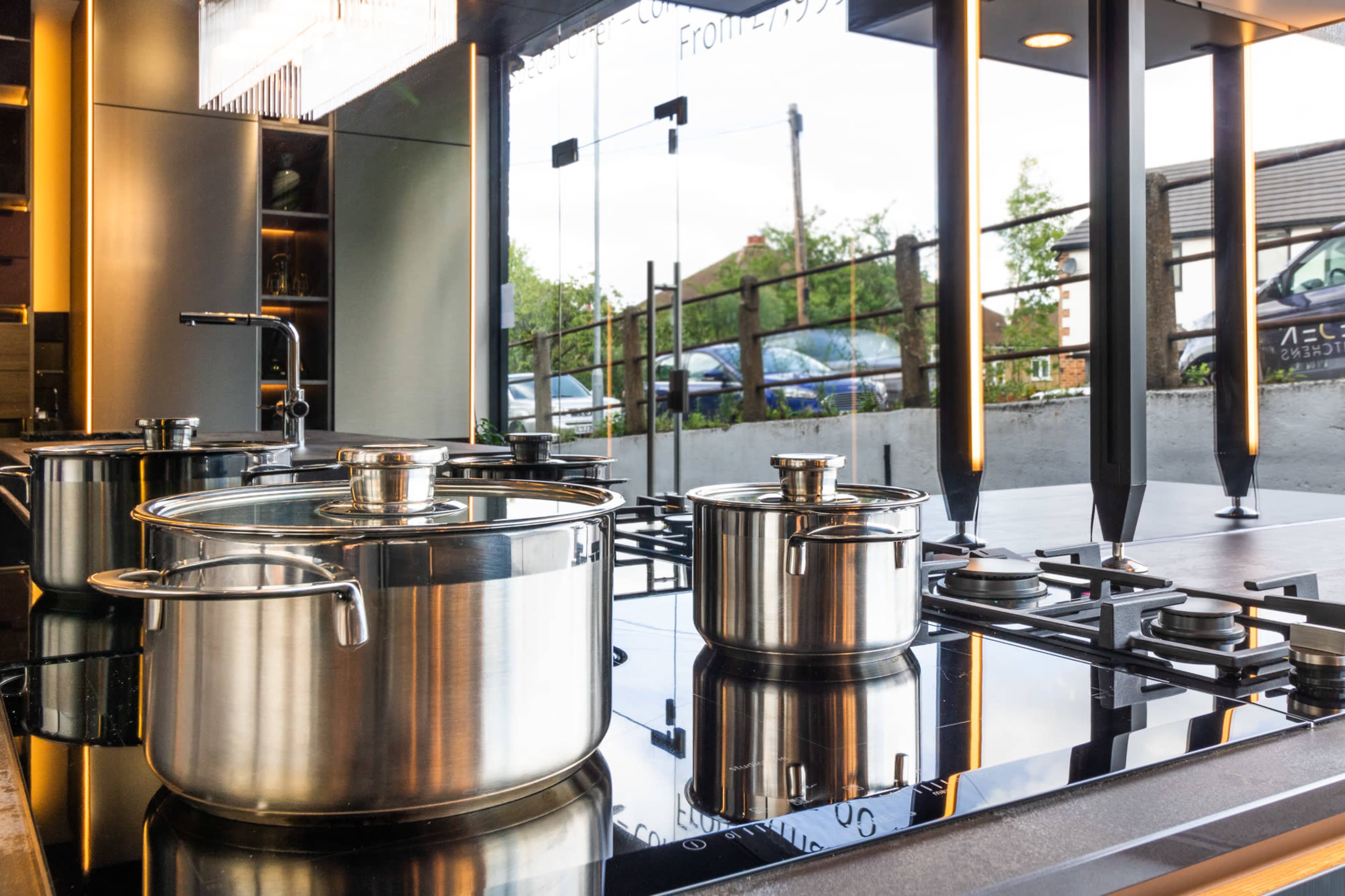 A modern kitchen with stainless steel pots on a glossy cooktop and large windows showing a view of parked cars outside.