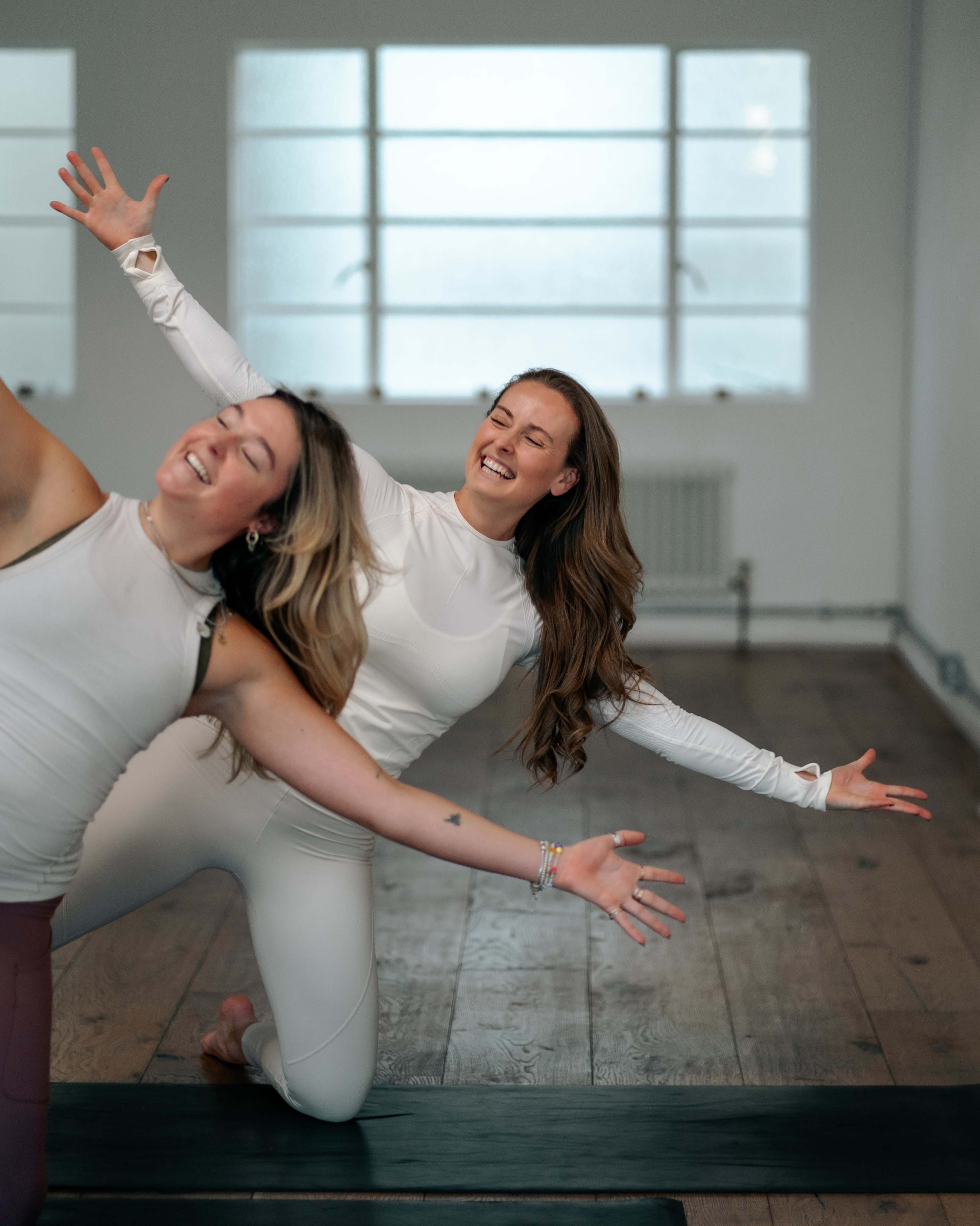 Two women in matching white outfits are joyfully posing on a yoga mat in a well-lit studio with large windows.