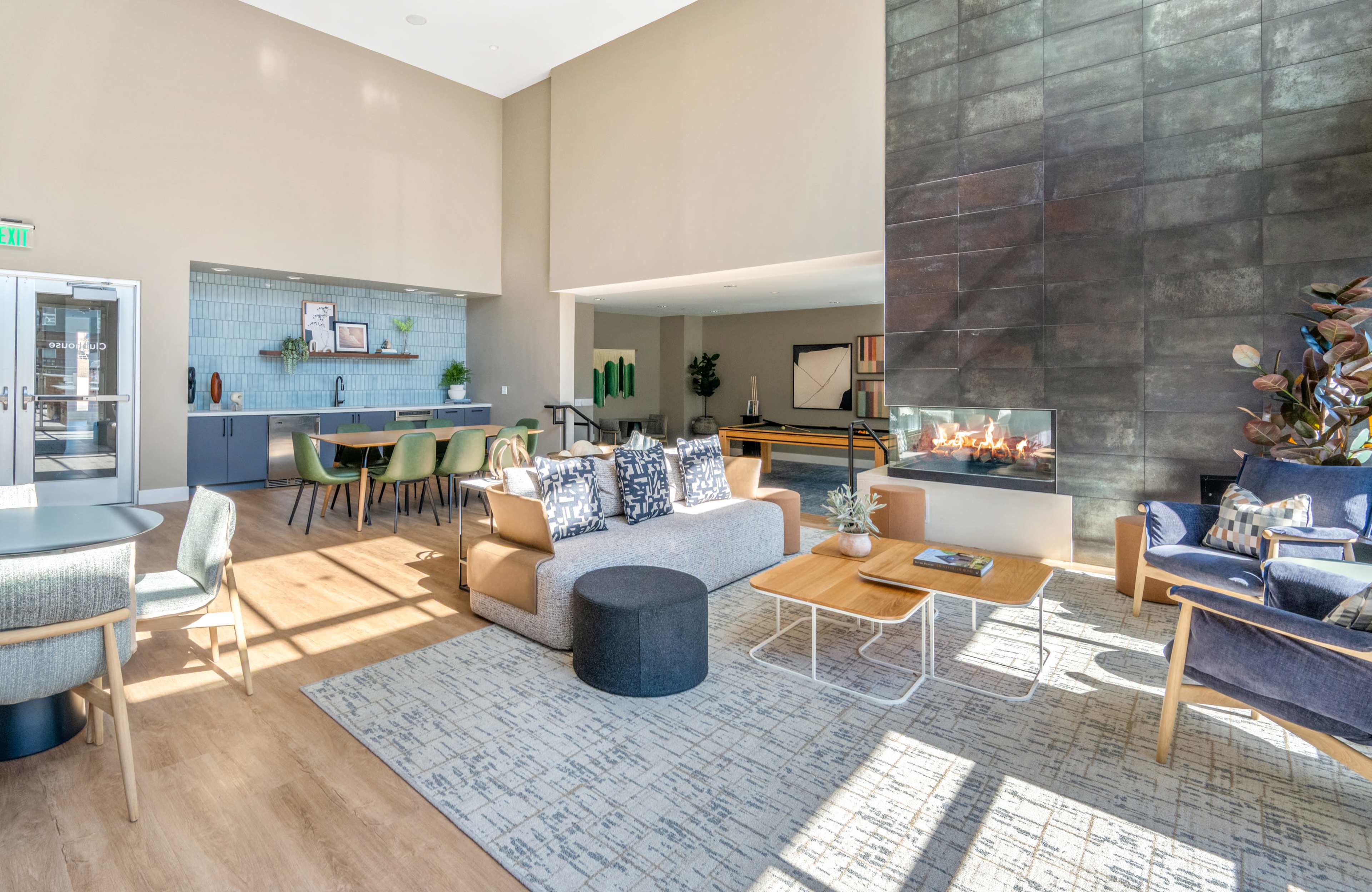 The image shows a modern lounge area with a fireplace, a mix of comfortable seating, and a kitchenette in the background.