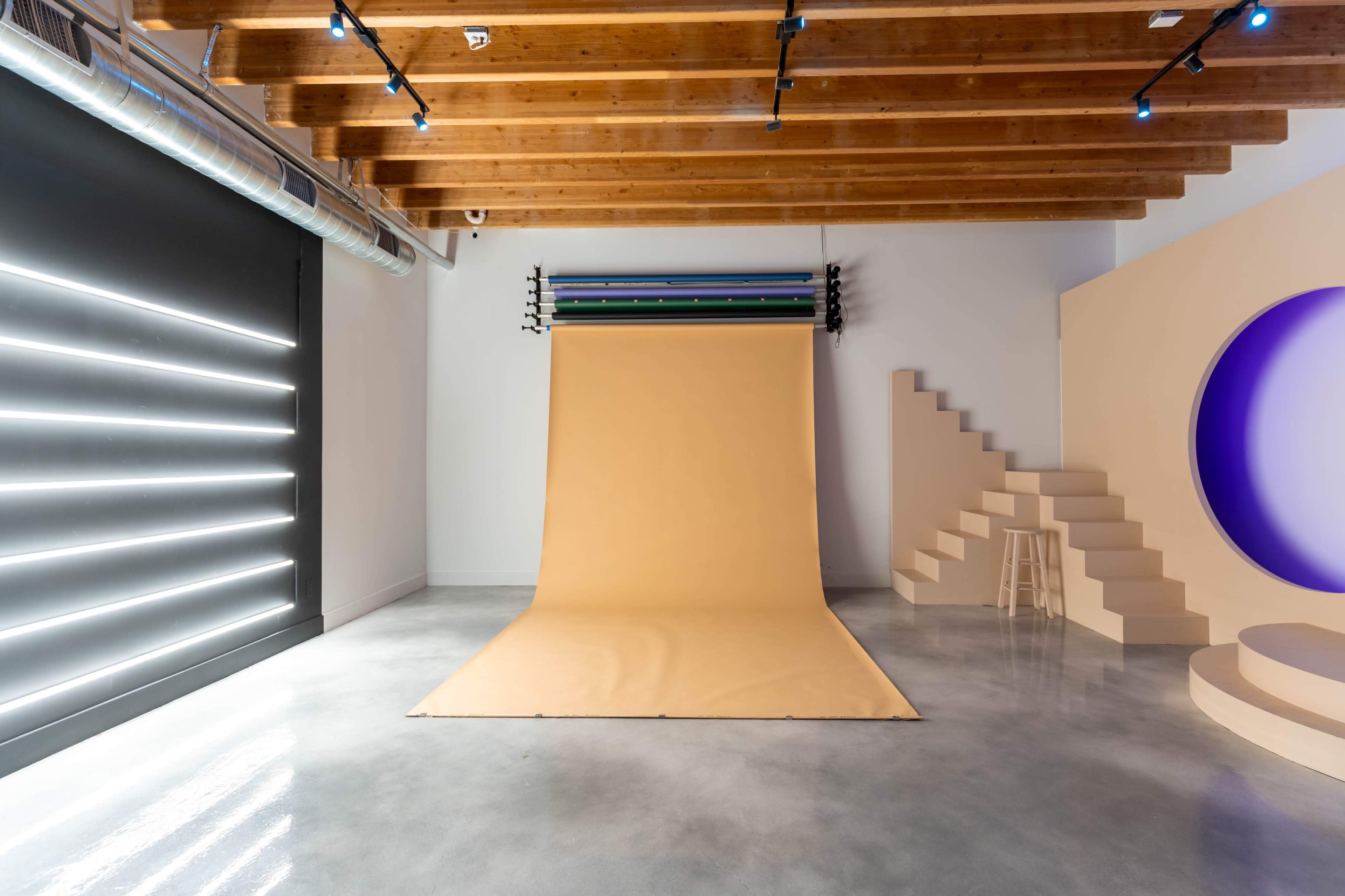 A spacious photo studio with a beige backdrop, wooden beams, and geometric elements, including a staircase and a circular cutout.